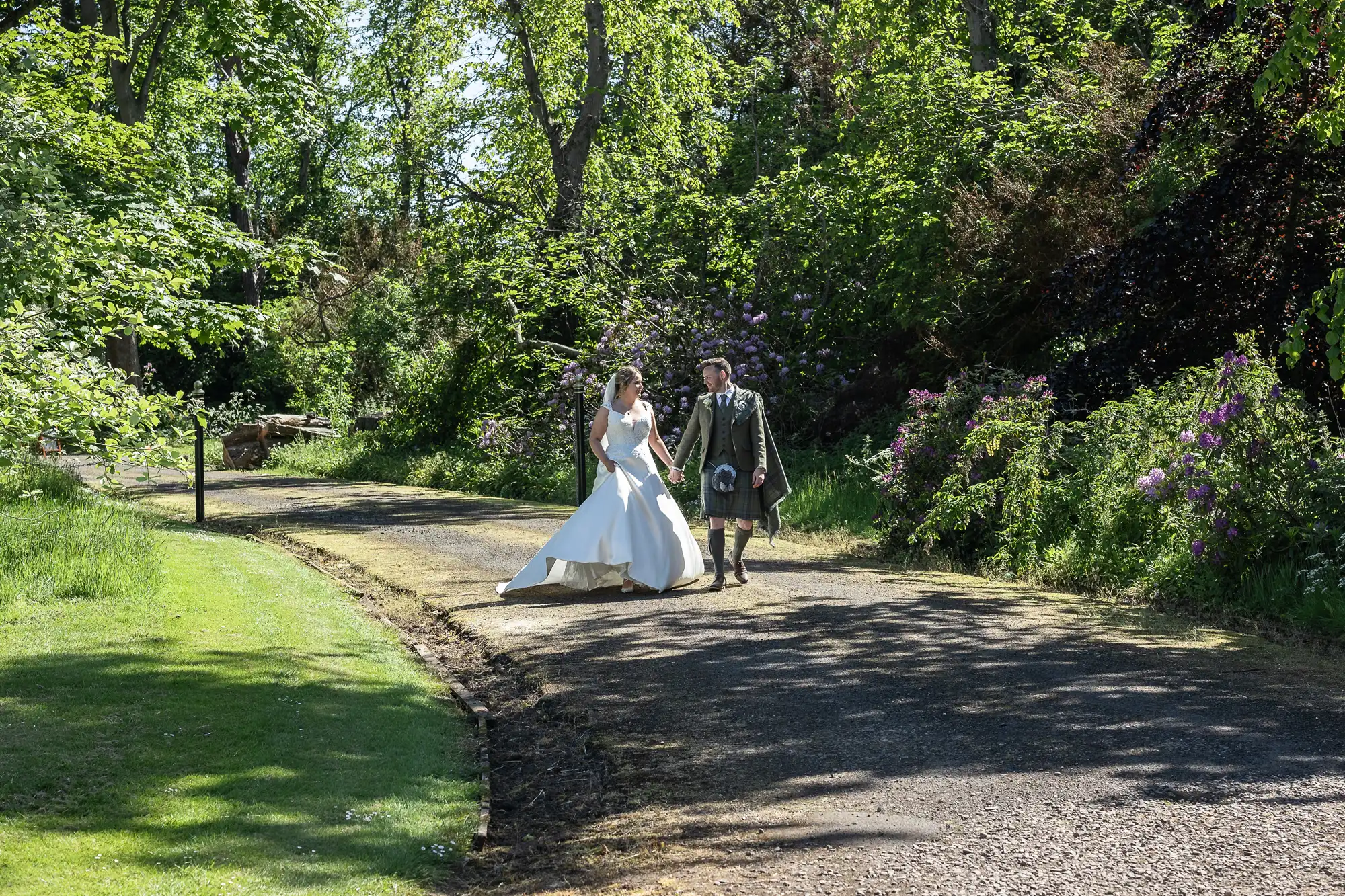 A bride in a white dress walks beside a groom in traditional Scottish attire along a tree-lined path on a sunny day.