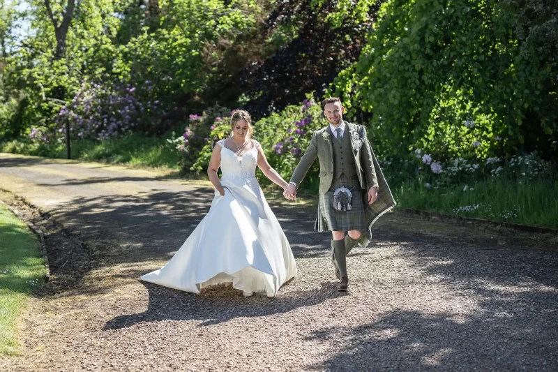 Netherbyres House wedding photos: a bride in a white gown and a groom in traditional Scottish attire walk hand-in-hand along a sunlit path surrounded by greenery.