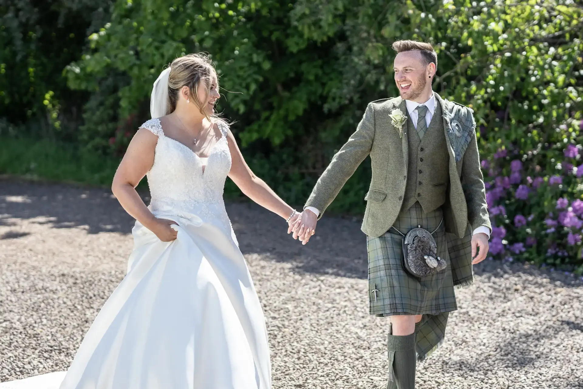 Netherbyres House wedding photographer image of a bride in a white dress and groom in a green kilt walk hand-in-hand outdoors, both smiling. Greenery and purple flowers are in the background.