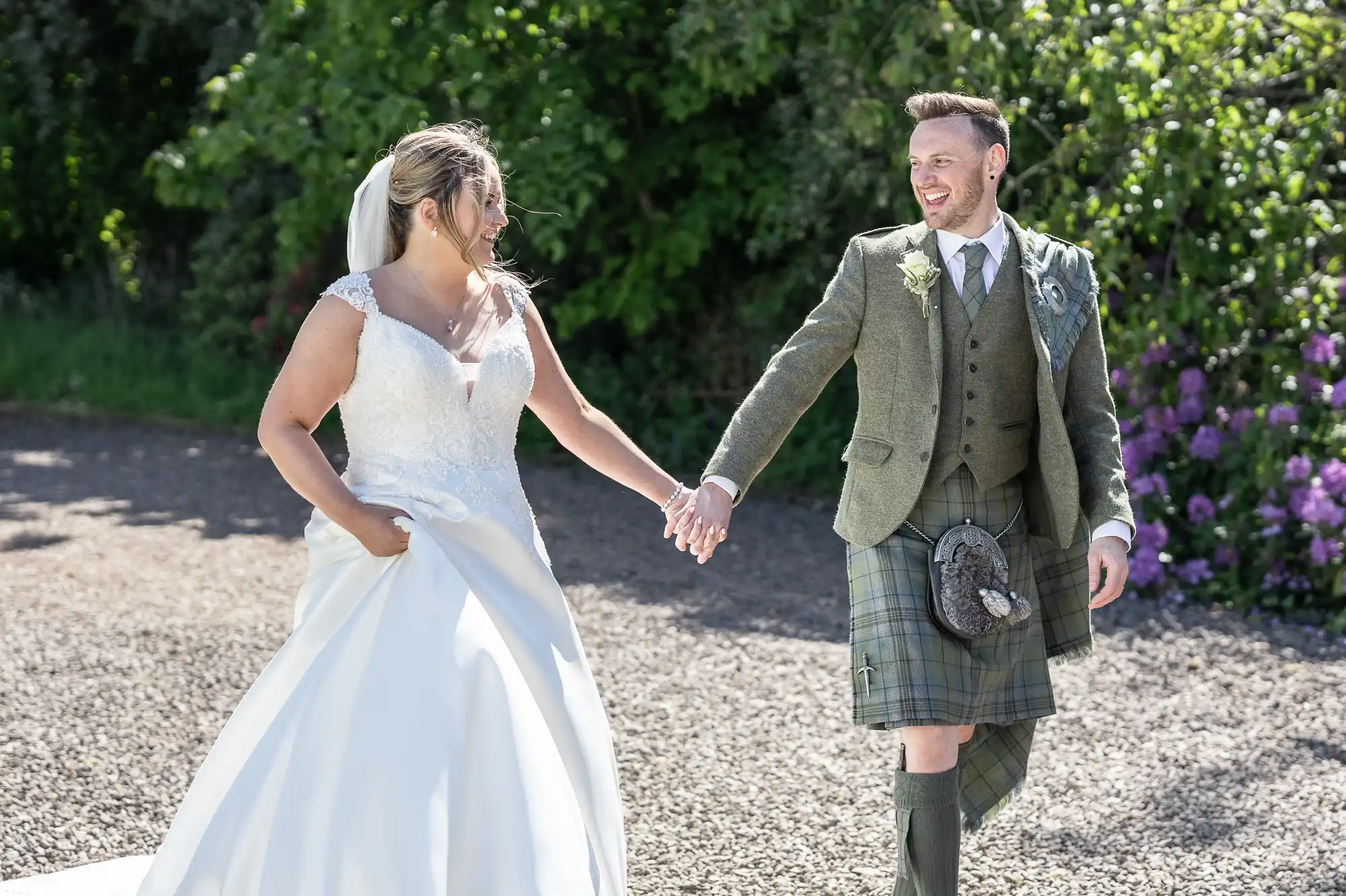 Bride in a white dress and groom in a green kilt walk hand-in-hand outdoors, both smiling. Greenery and purple flowers are in the background.