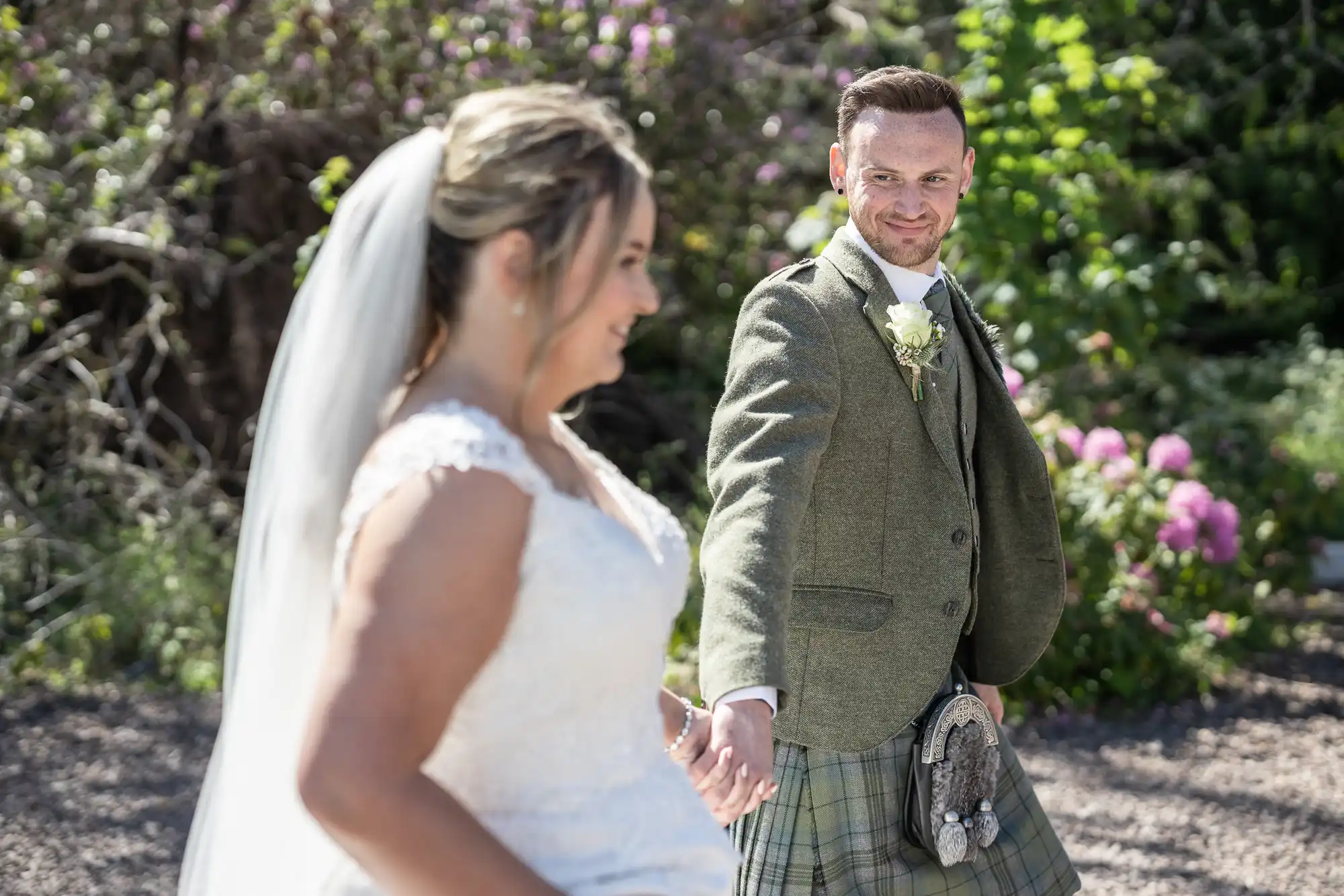 A bride in a white gown and veil walks hand in hand with a groom in a green tweed jacket and kilt. They are outdoors with greenery and flowers in the background.