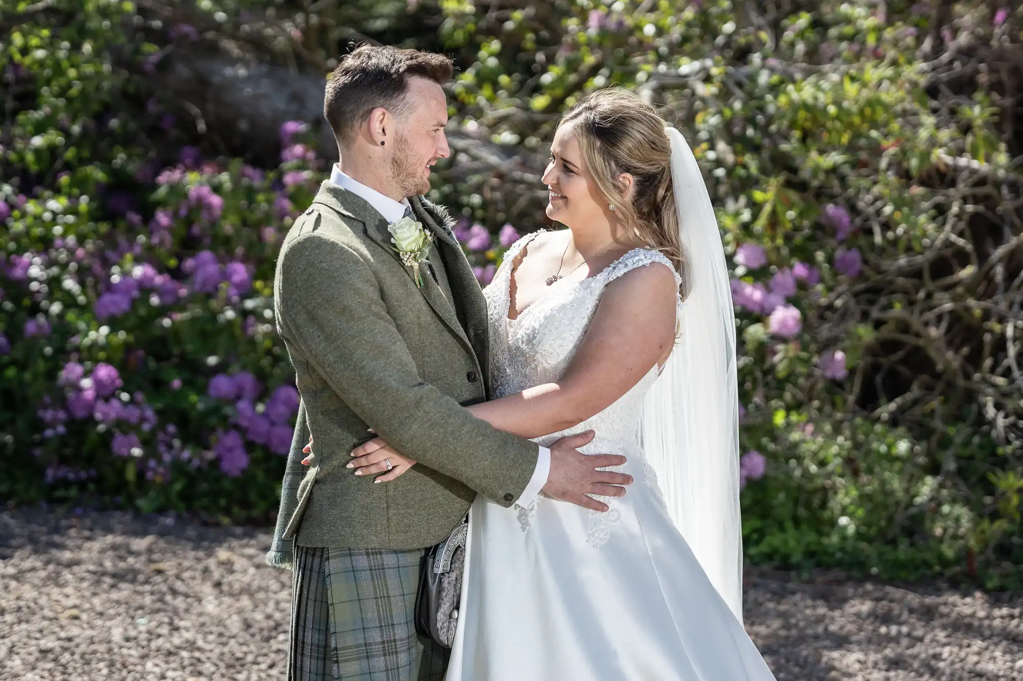 A bride and groom look at each other and smile while embracing. The bride wears a white dress and veil; the groom is in a green suit. They stand outdoors with greenery and flowers in the background.