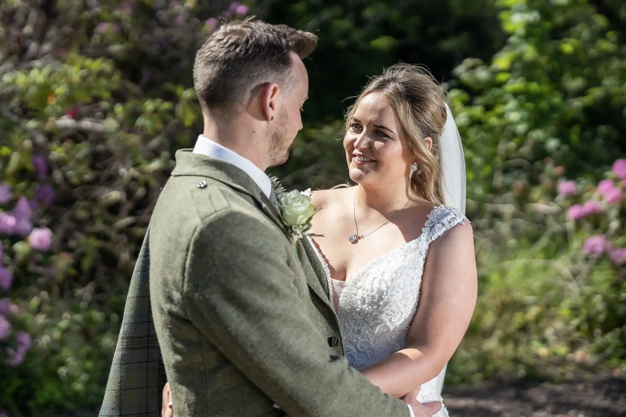 A bride and groom in wedding attire stand close, smiling at each other. The bride wears a white dress with lace detail; the groom is in a green suit with a boutonniere. Trees and flowers are in the background.