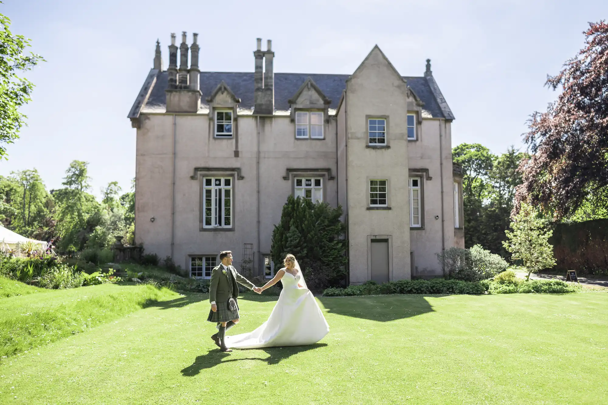 A couple, with the groom in a kilt and the bride in a white gown, hold hands on a green lawn in front of a large historic building.