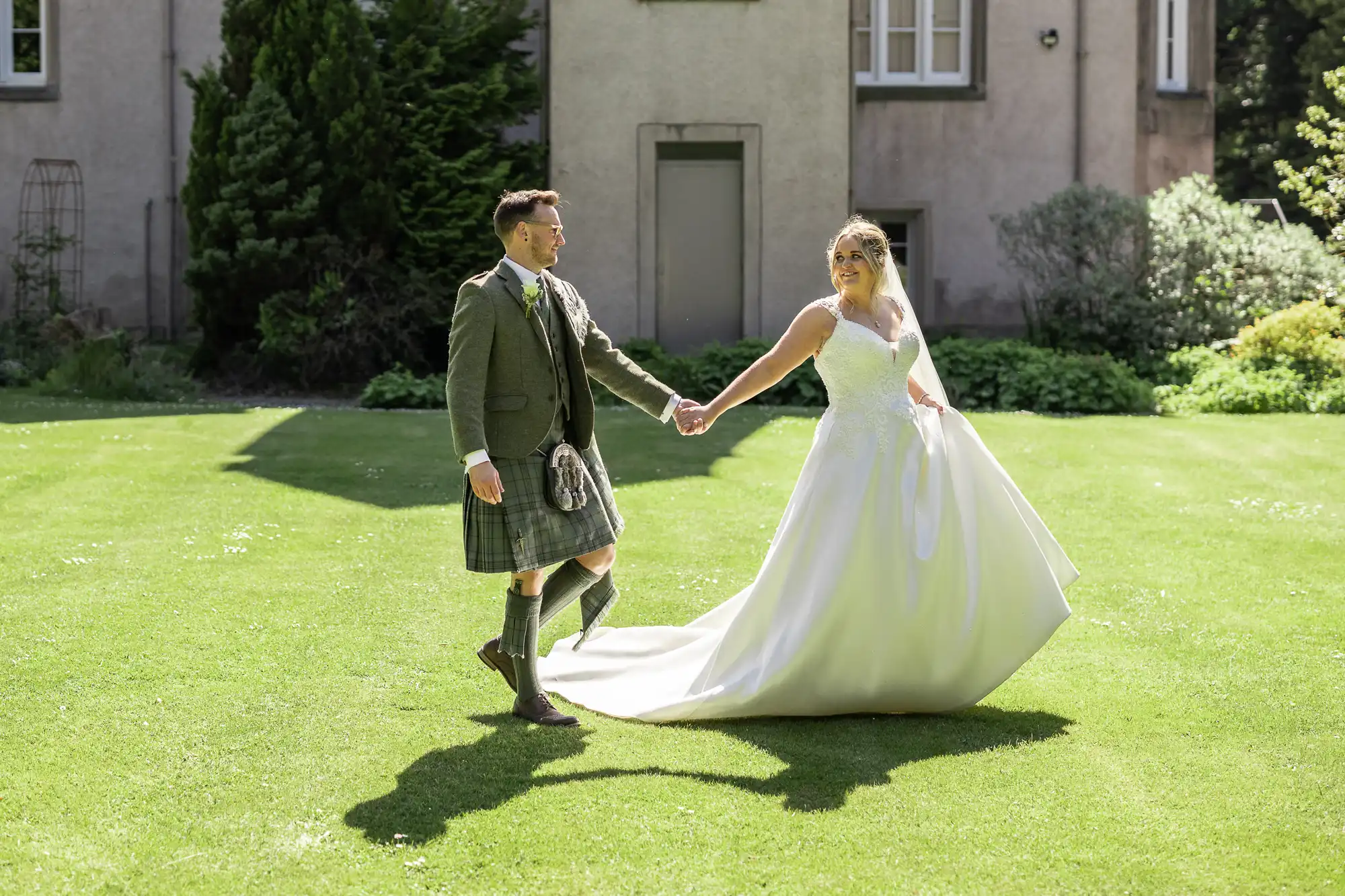 A couple dressed in wedding attire holds hands and walks on a lawn near a building. The bride wears a white gown, and the groom wears a kilt.