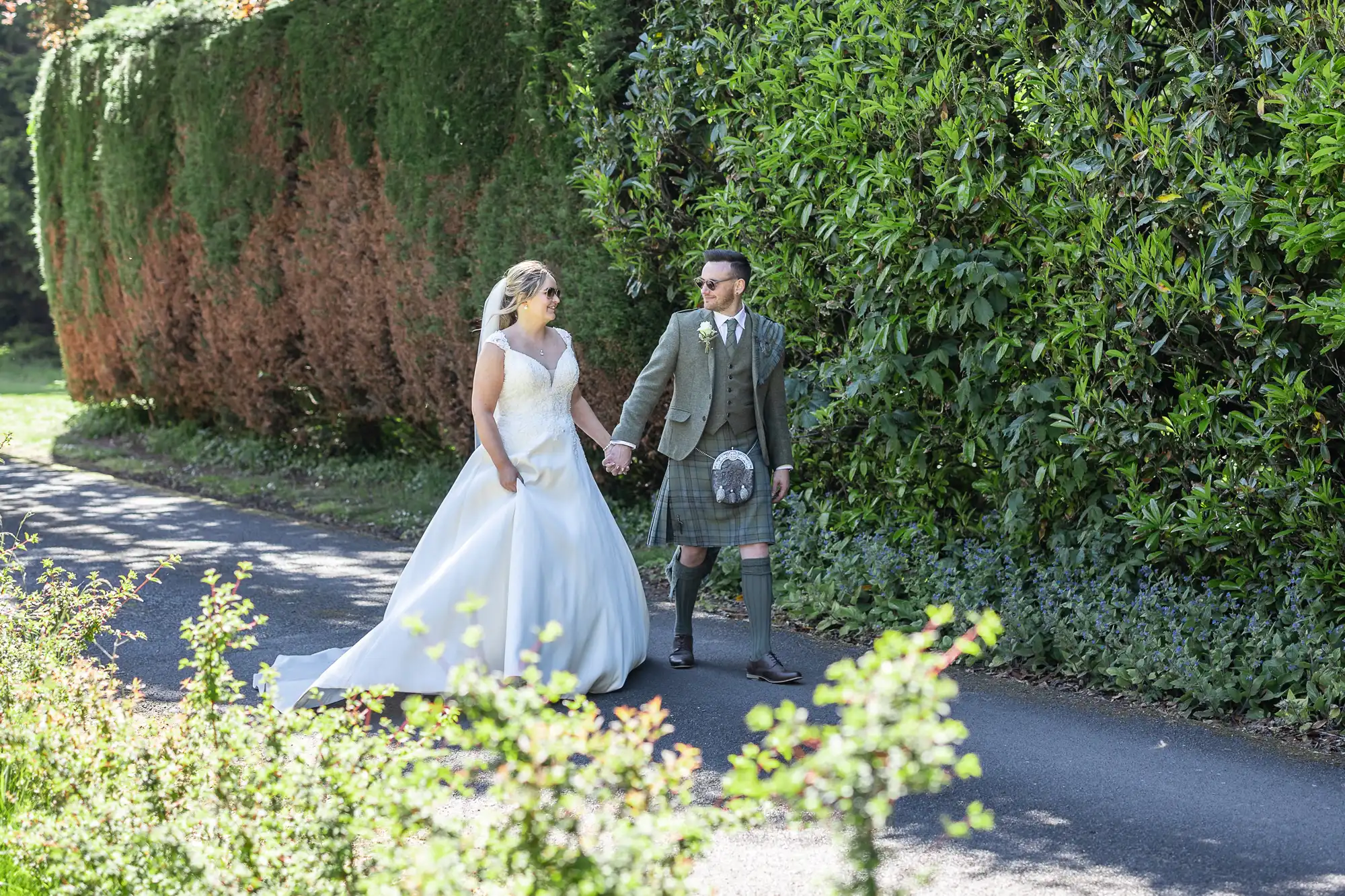 A bride in a white gown and a groom in traditional Scottish attire walk hand in hand on a pathway, surrounded by greenery.