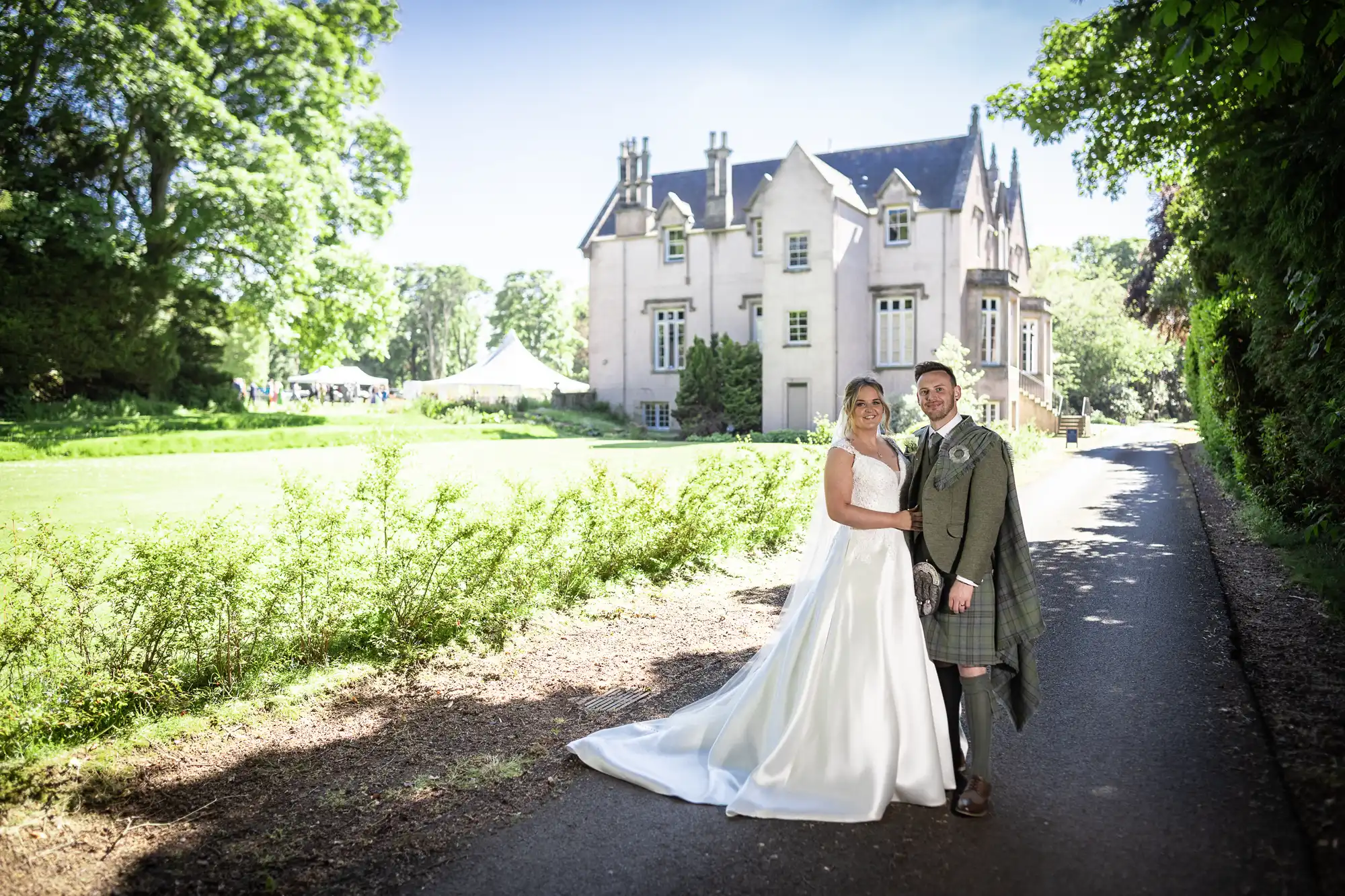 A couple in wedding attire, the groom in a kilt and the bride in a white dress, pose on a tree-lined path in front of a large, historic building.