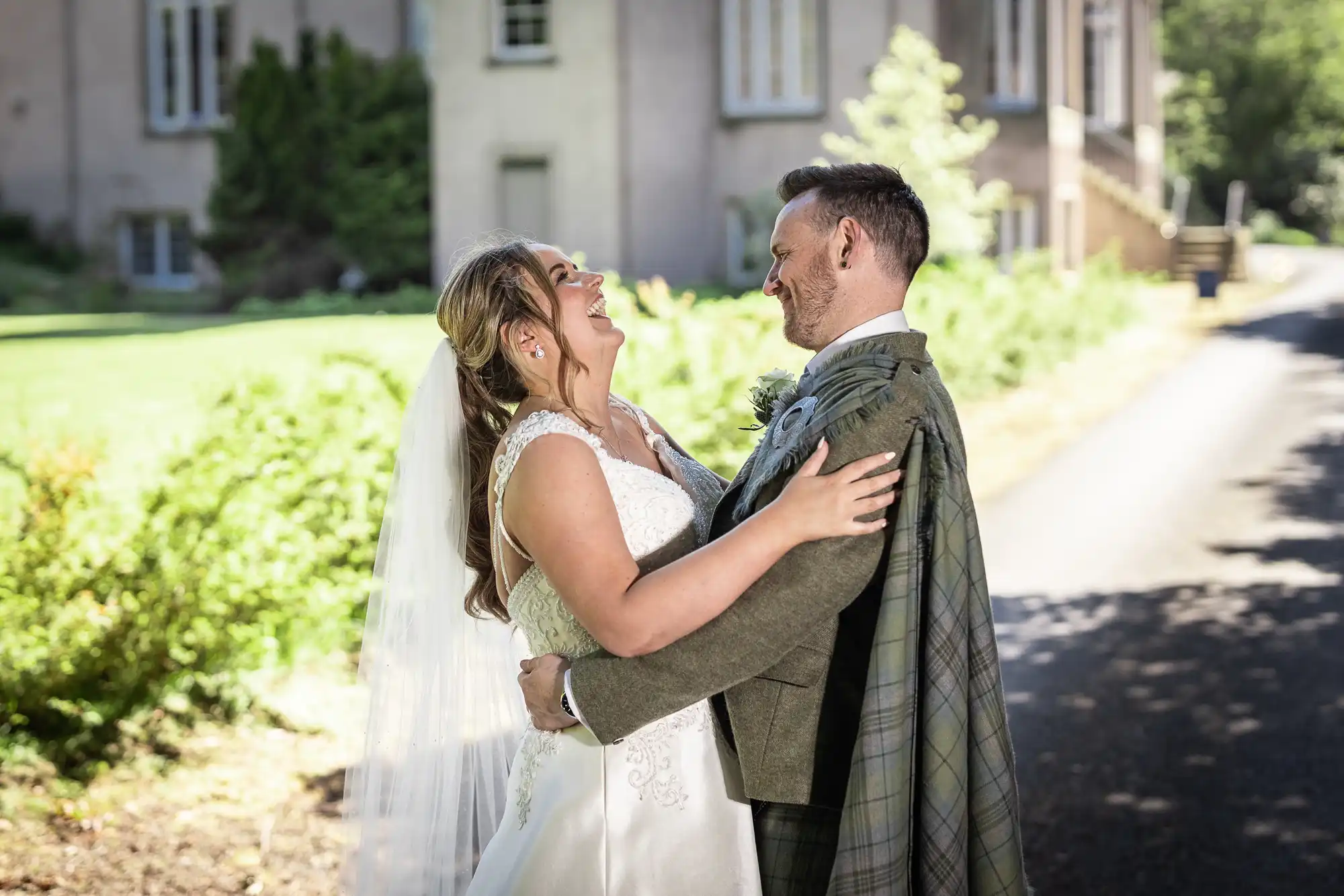 A bride in a white dress and veil and a groom in a greenish suit and tartan attire share a joyful moment outdoors, gazing into each other's eyes and smiling.