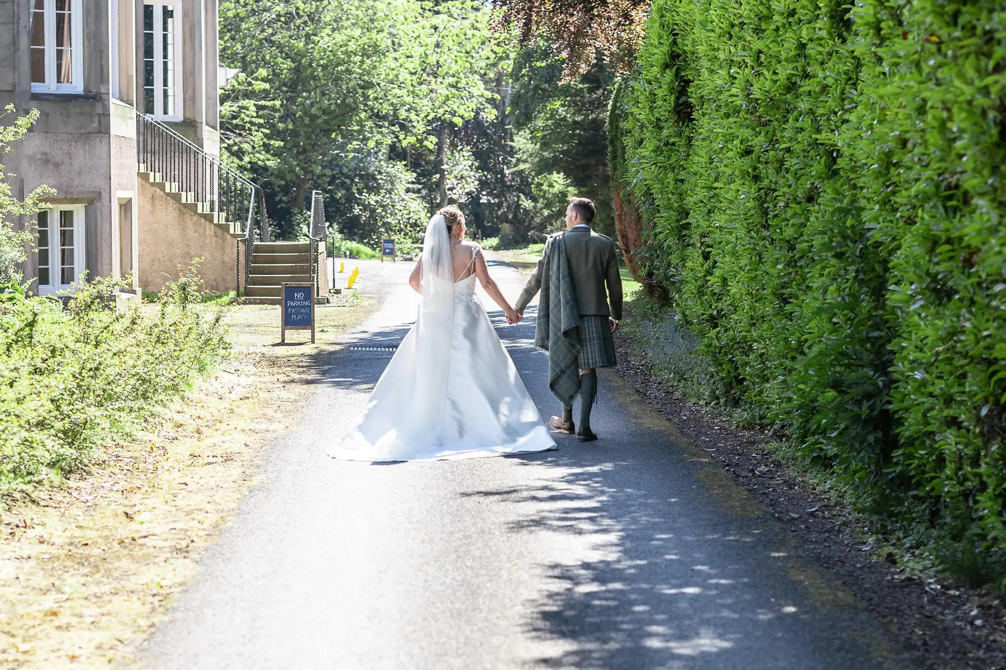 A bride and groom holding hands walk down a sunlit path flanked by green hedges and a building on the left.