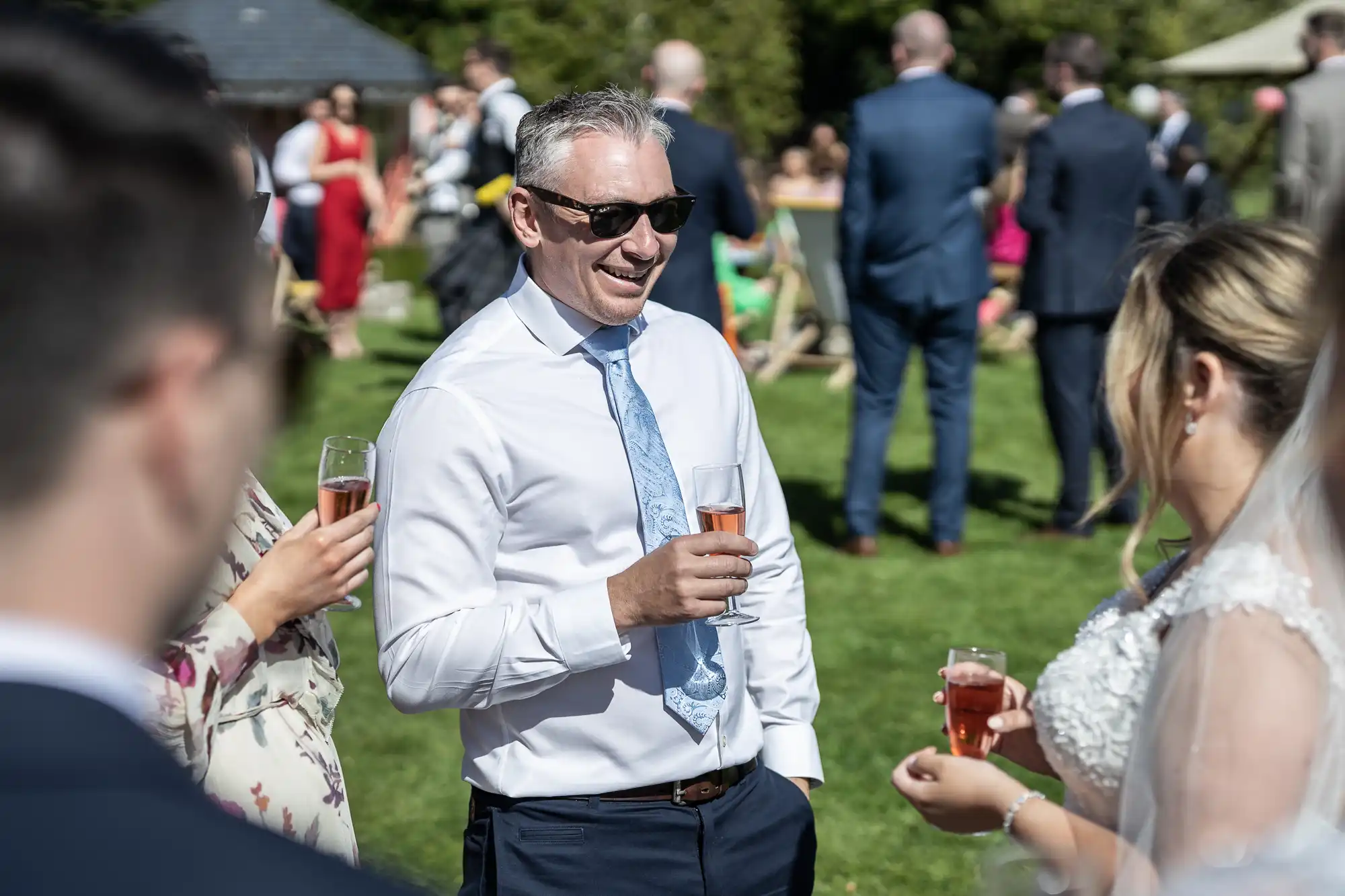 A man in a white shirt and sunglasses holds a drink while conversing at an outdoor daytime event. Several people are gathered in the background.