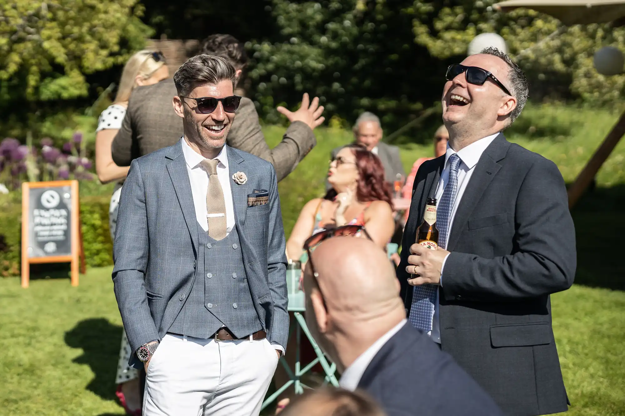 Two men in suits, one wearing sunglasses, are laughing and talking at an outdoor event. Other guests can be seen in the background.