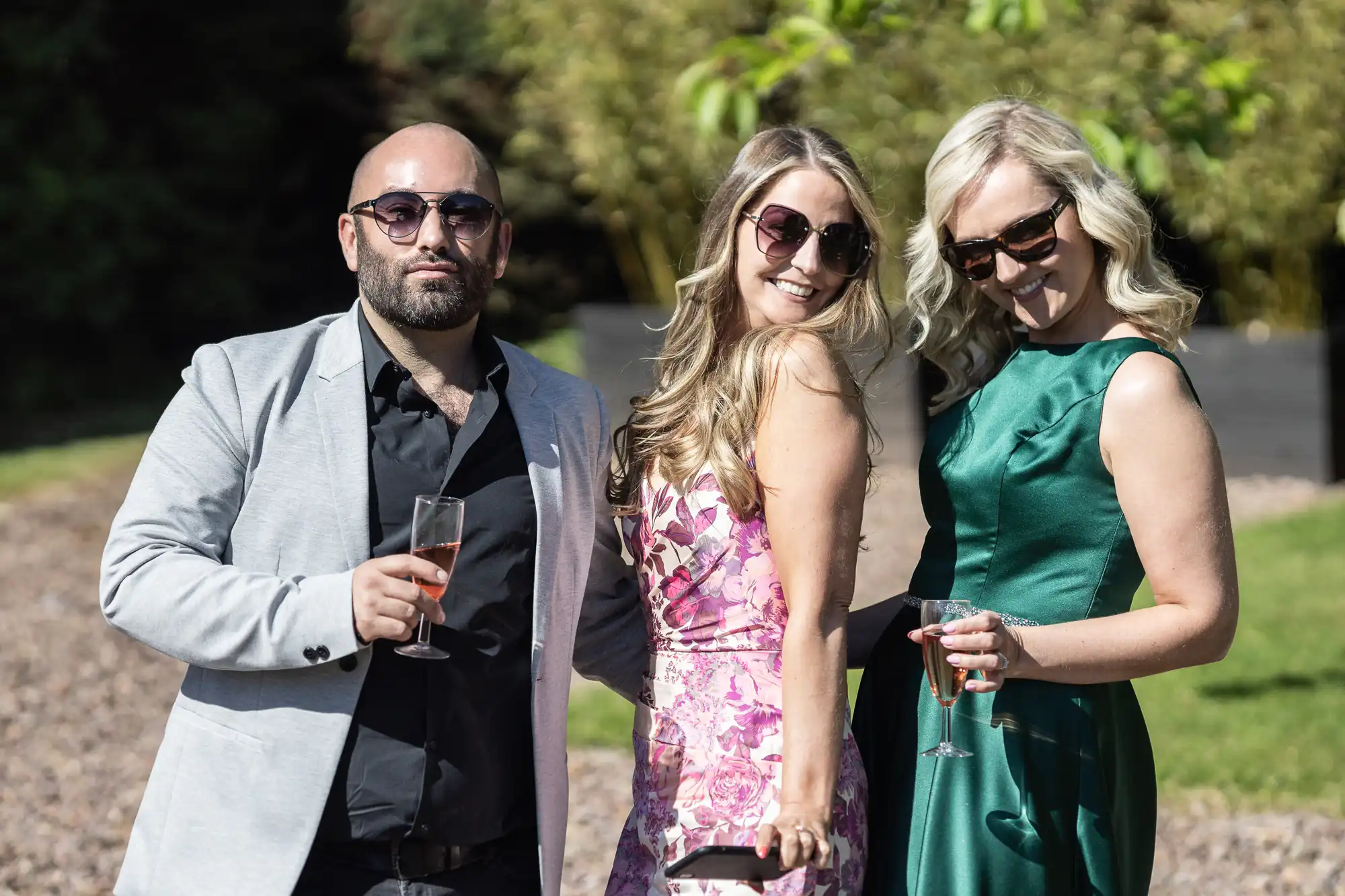 A man in a gray blazer and two women in dresses, both wearing sunglasses, smile while holding drinks outside on a sunny day.