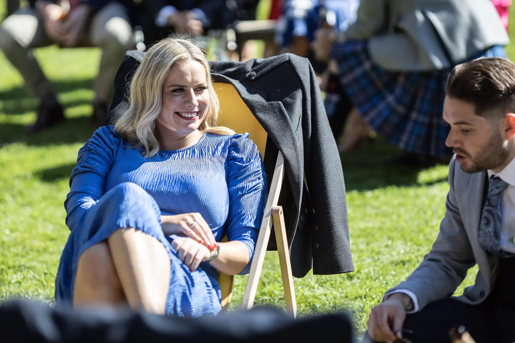 Woman in a blue dress sitting on a deck chair, talking to a man in a suit outdoors on a sunny day with other people in the background.