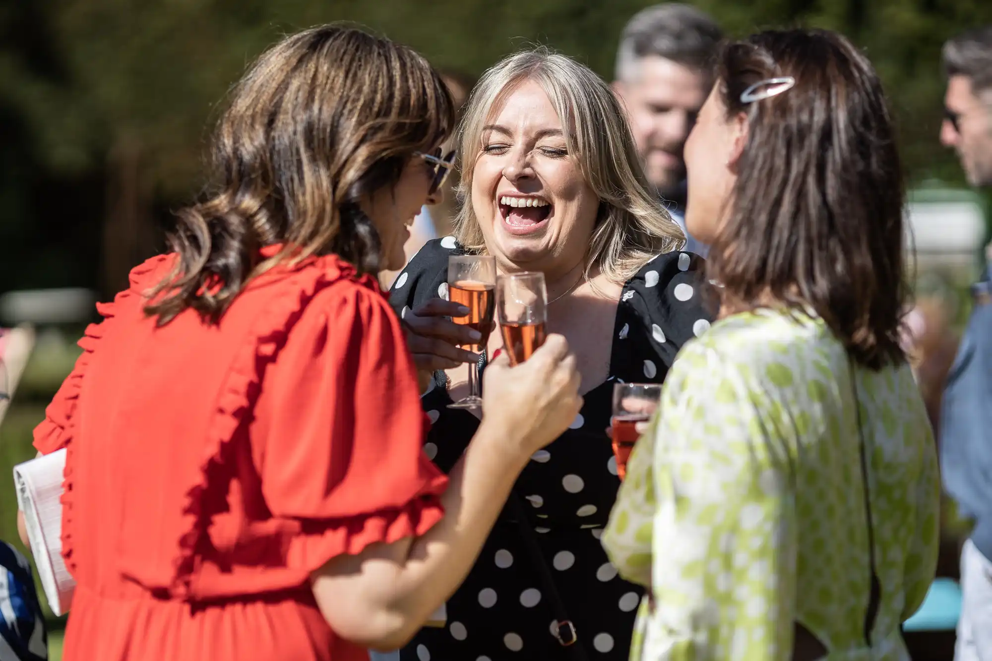 Three women stand outside, holding glasses of pink drink, laughing and smiling. The woman in the center wears a black and white polka dot dress. Other people are blurred in the background.