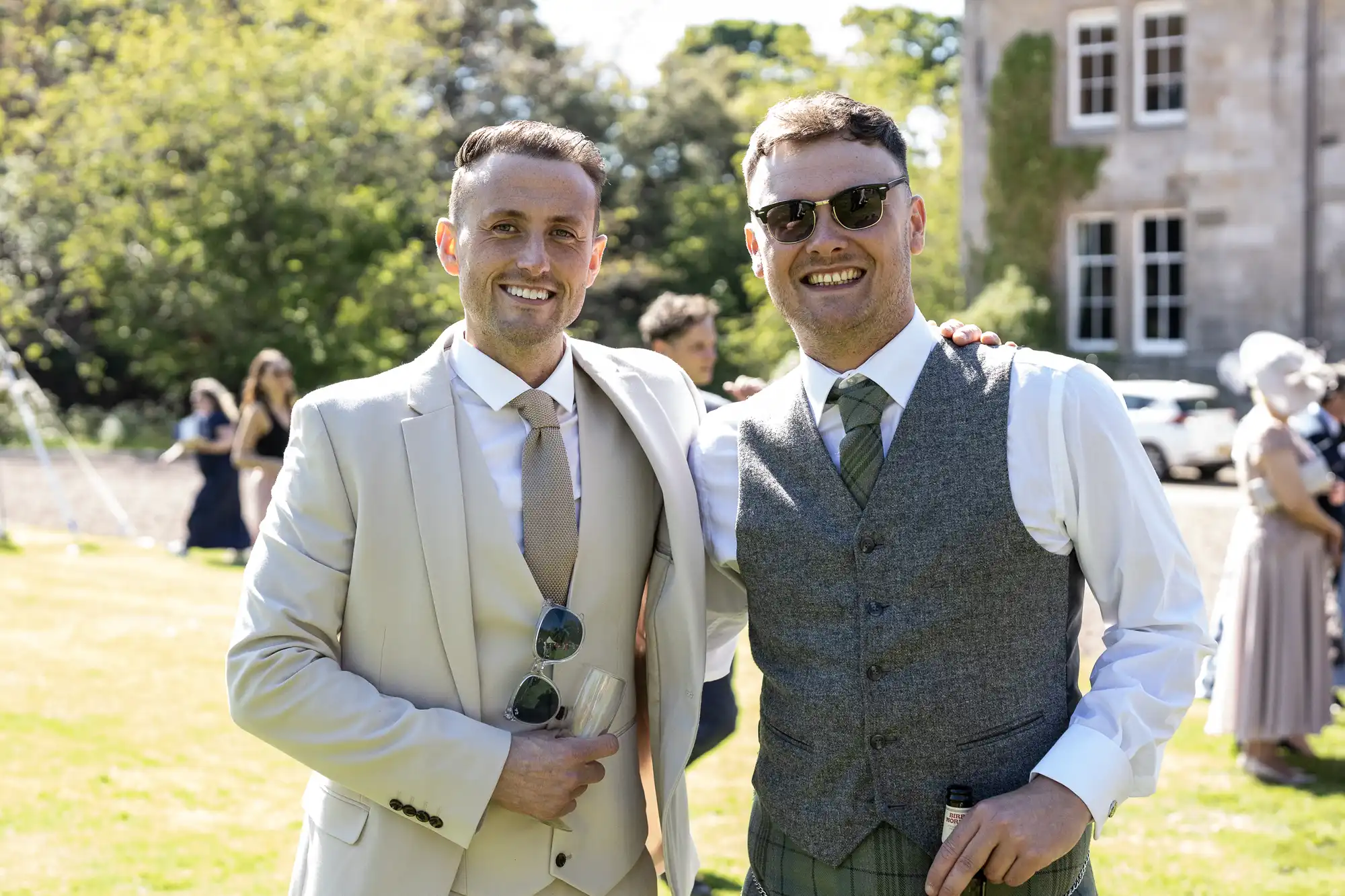 Two men dressed in suits stand outdoors at a social event, smiling at the camera. The man on the left wears a beige suit, and the man on the right wears a grey vest and sunglasses.