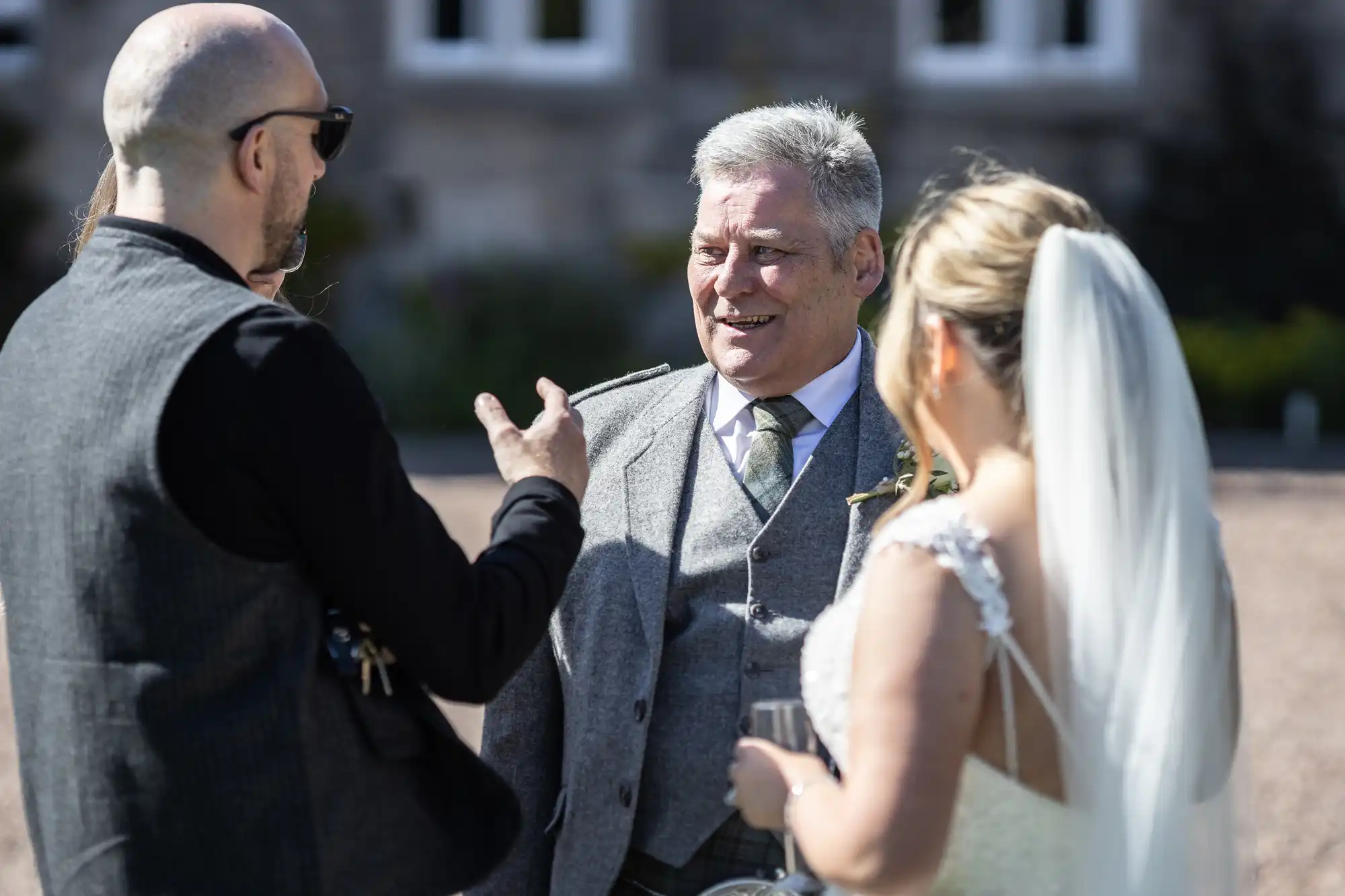 Three people are standing outdoors, engaged in conversation. The man in the middle is smiling, and the woman on the right appears to be a bride wearing a white dress and veil.