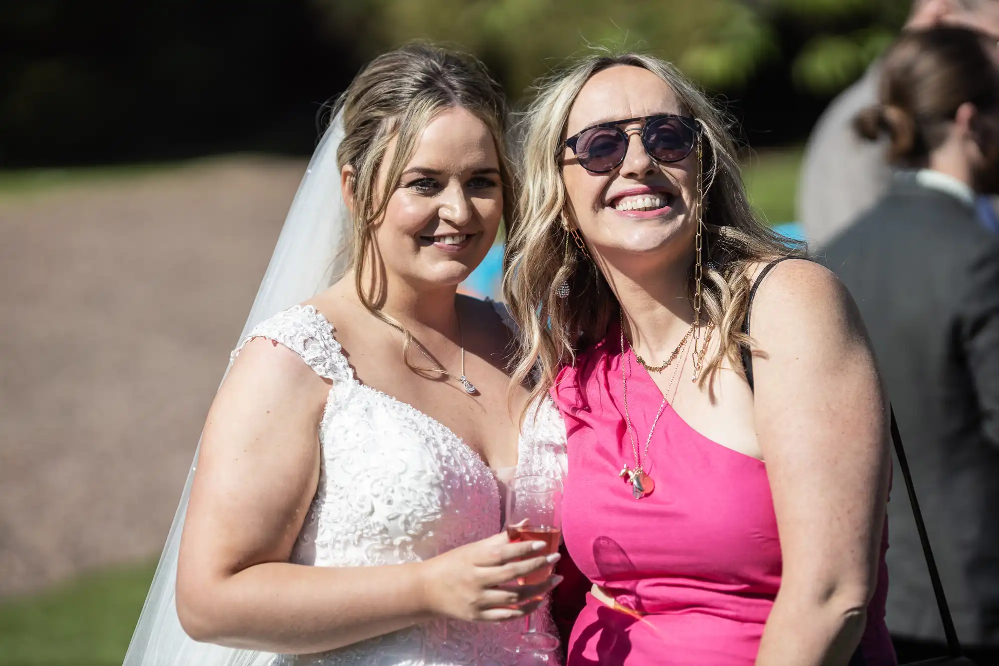 A bride in a white dress holding a drink stands next to a woman in a pink dress and sunglasses at an outdoor event.