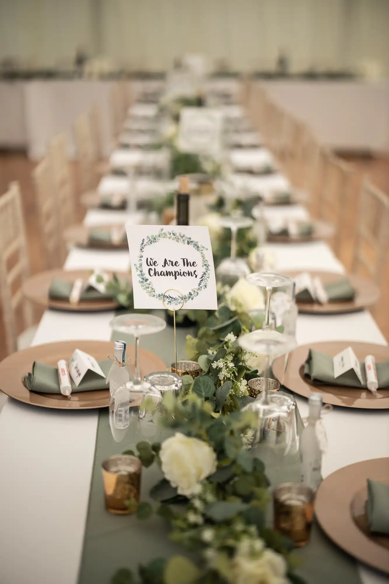 A long banquet table is set with plates, glassware, and greenery. A card at the center reads "We Are The Champions".