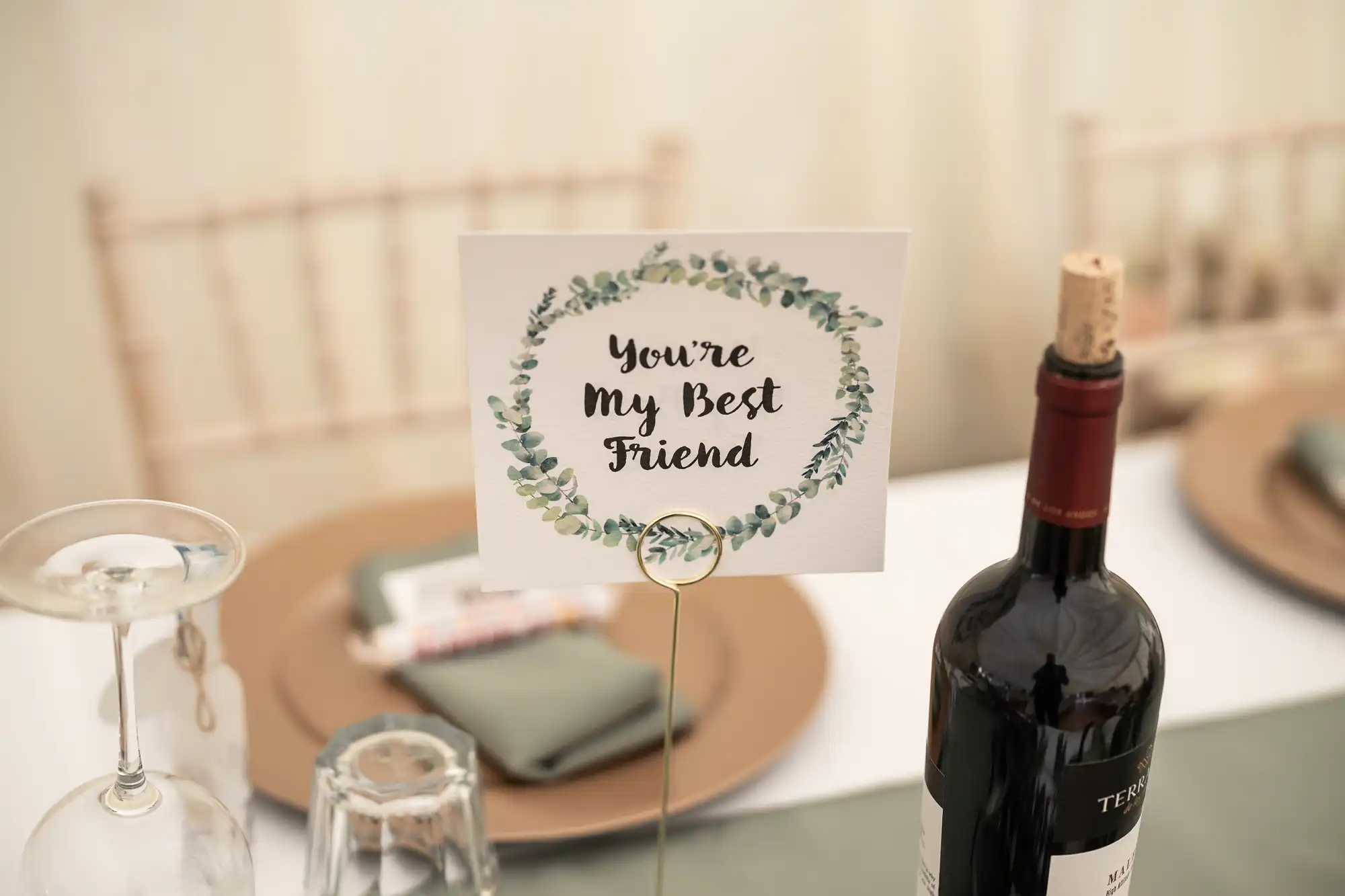 A table setting with a wine bottle, an empty wine glass, and a sign that reads "You're My Best Friend." The backgrounds include empty chairs and neatly arranged plates and napkins.