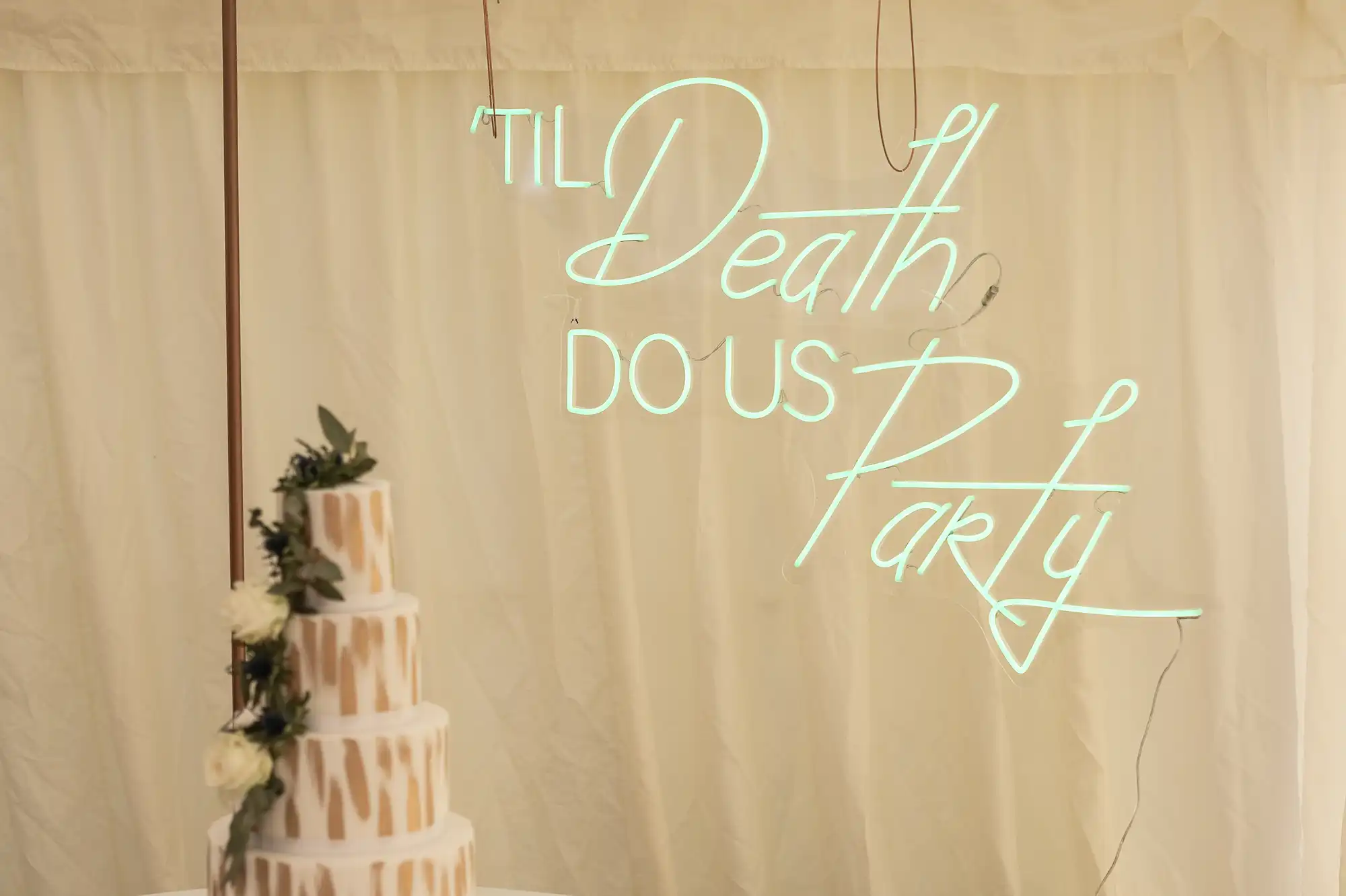 A three-tiered wedding cake with white icing and green foliage decoration is in front of a neon sign that reads "'Til Death Do Us Party.