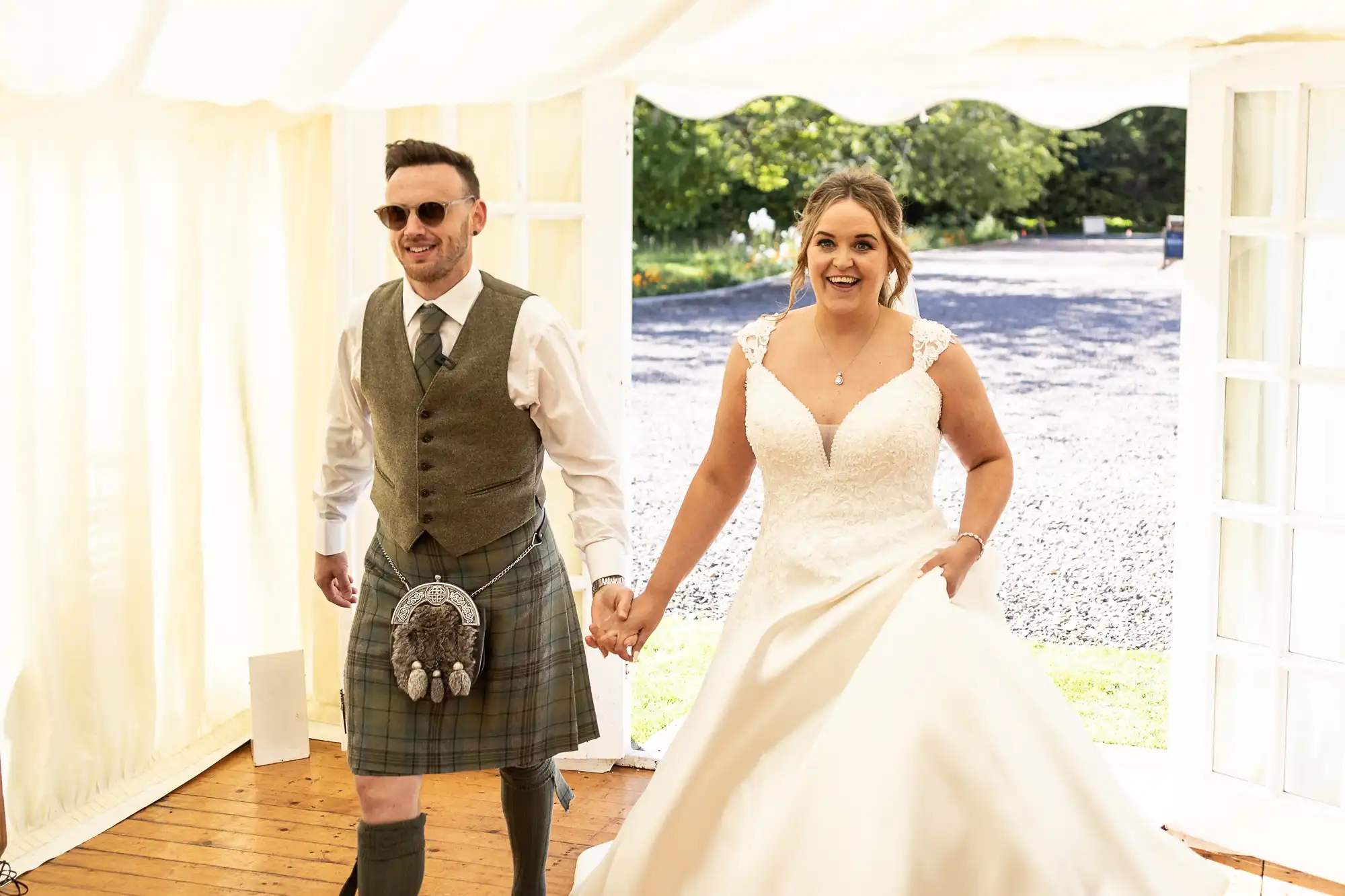 A man in a kilt and sunglasses holds hands with a woman in a white wedding dress as they walk inside a tent on a sunny day.