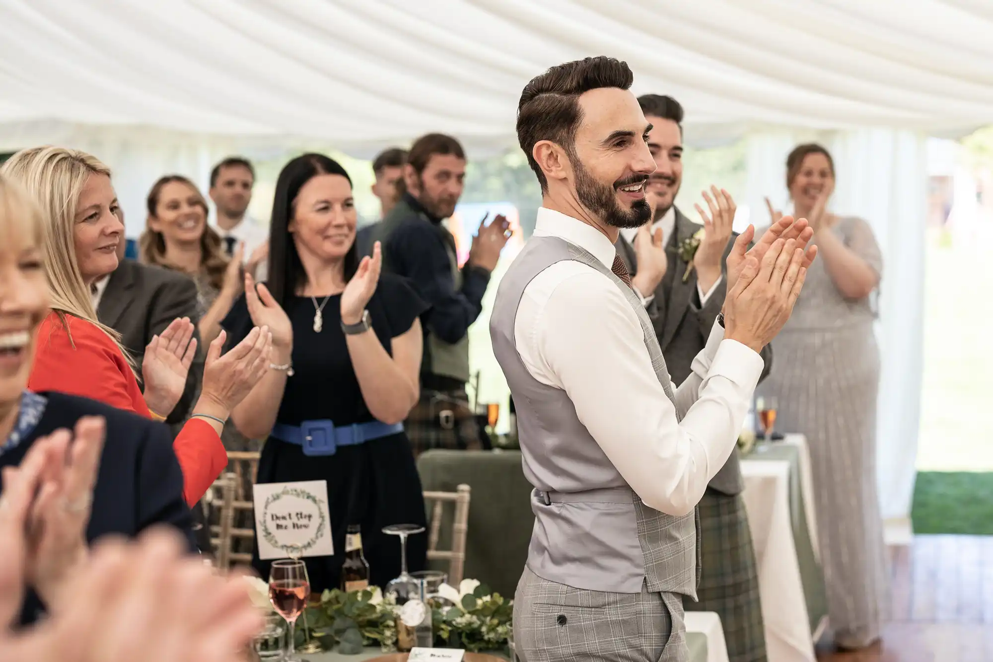 A man in a suit is clapping while standing under a white canopy, surrounded by a crowd of people who are also clapping, at what appears to be a celebratory event.