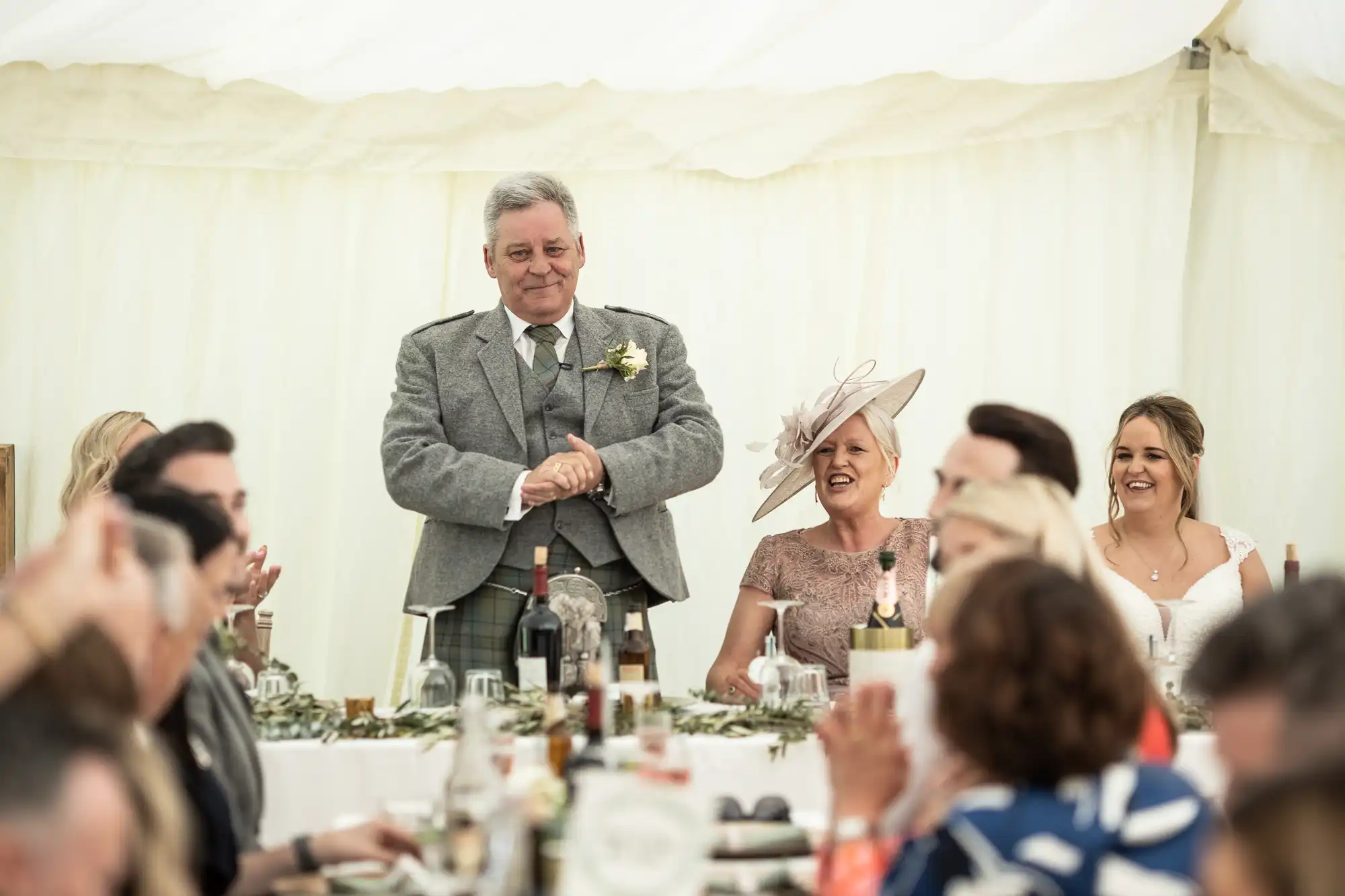 A man in a gray suit stands and smiles during an event, with seated guests clapping. Two women, wearing formal attire, sit to his right, and a decorated table is in the foreground.