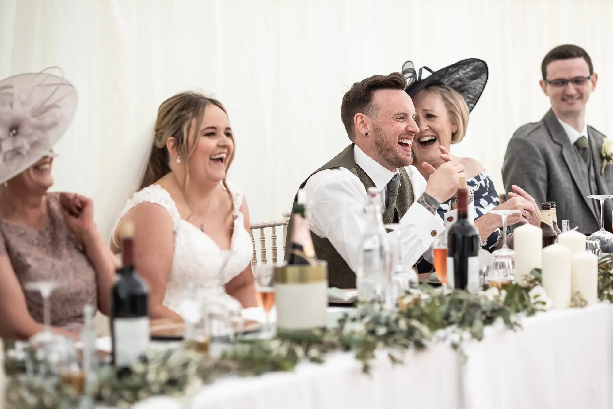 A wedding party laughs and smiles while seated at a long, decorated table. The bride, groom, and other guests are engaged in conversation and enjoying the celebration.