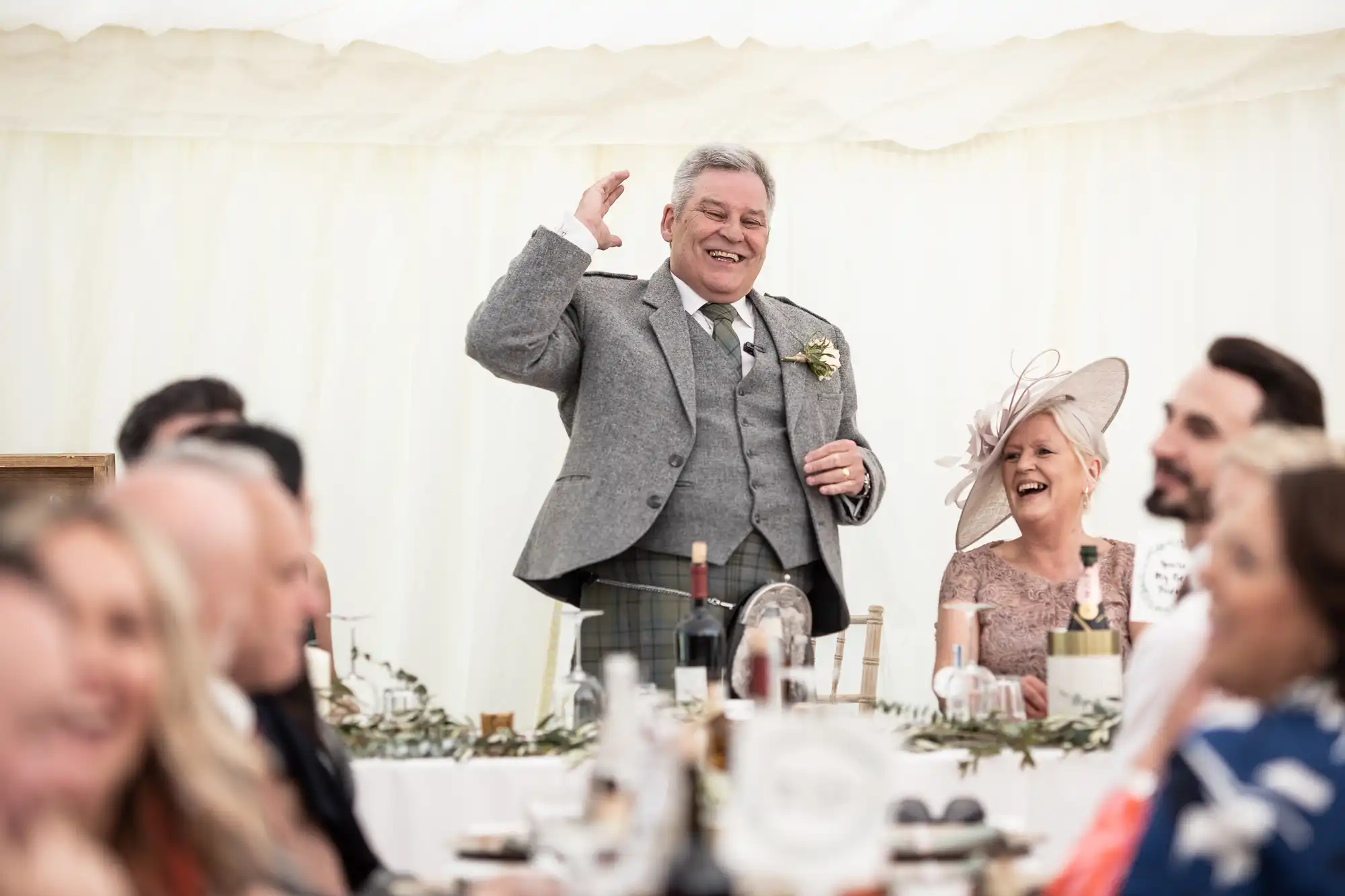 An older man, dressed in a grey suit and tie, stands and waves, smiling at a wedding reception. Seated guests watch and smile, with bottles and glasses on the table in front of them.