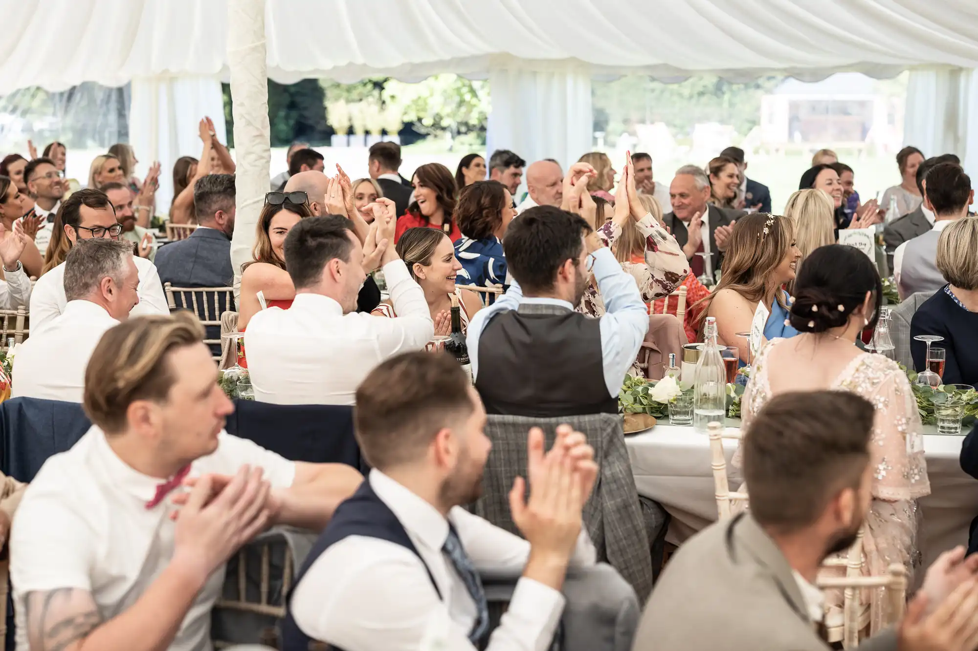 A large group of people seated at tables under a white canopy, applauding and engaging with each other during a formal event.