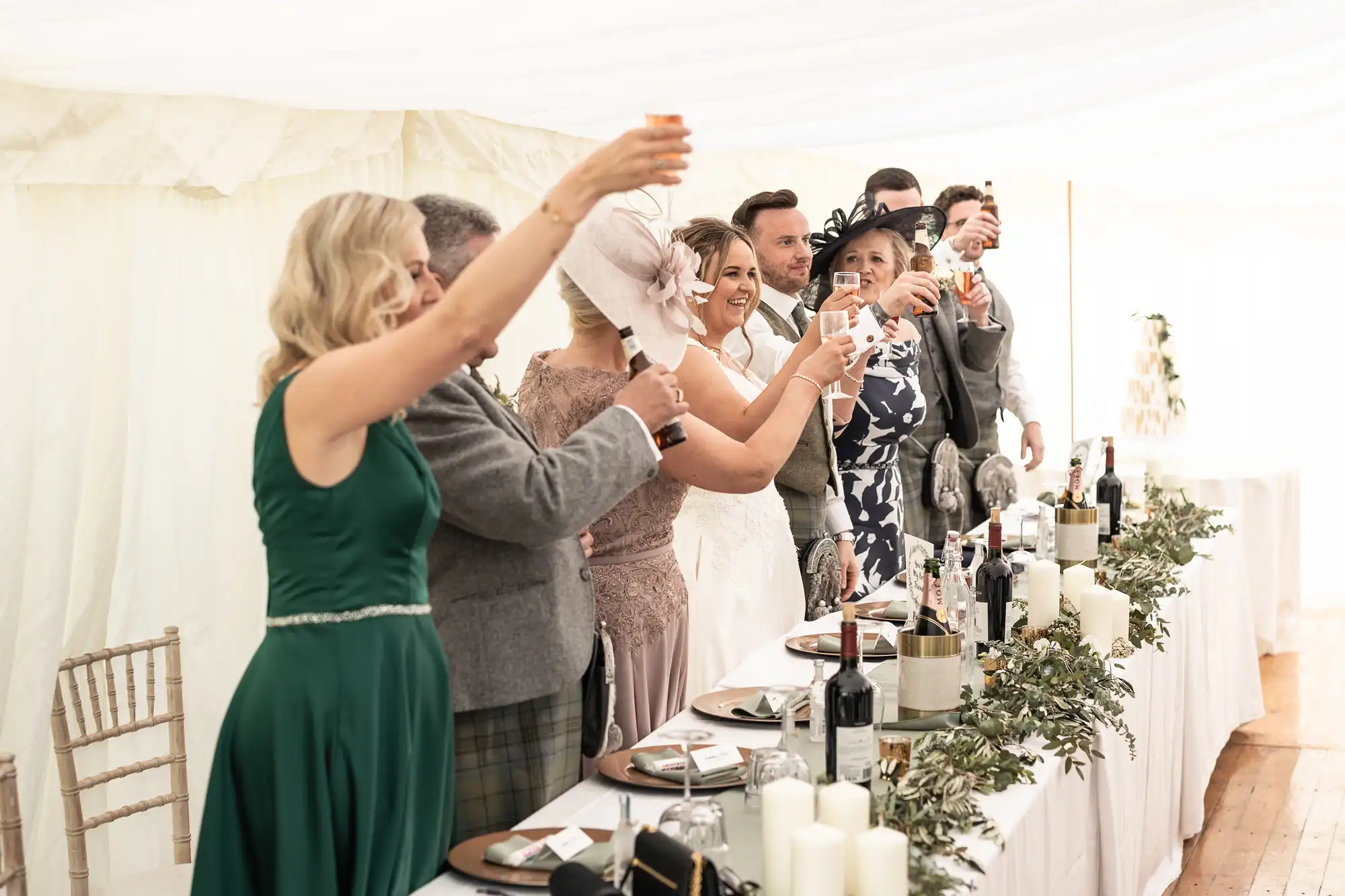 A group of people, including a bride and groom, raise their glasses in a toast at a wedding reception. The table is set with wine bottles, greenery, and candlesticks.