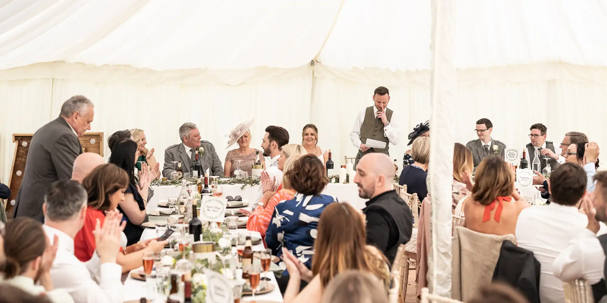 A wedding reception in a tent with guests seated at tables while a man stands and speaks into a microphone near the bridal party.