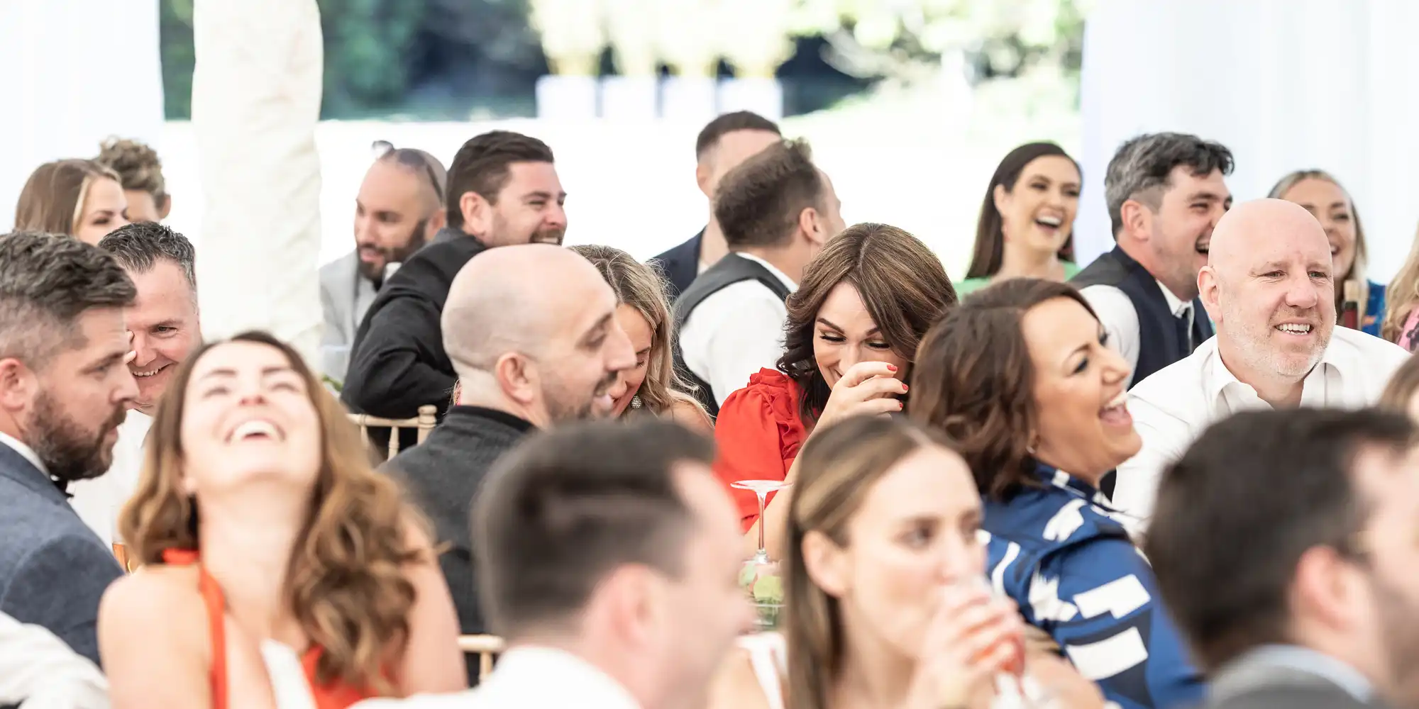 A large group of people, dressed in various formal attires, sit closely together at an indoor event, smiling and laughing.