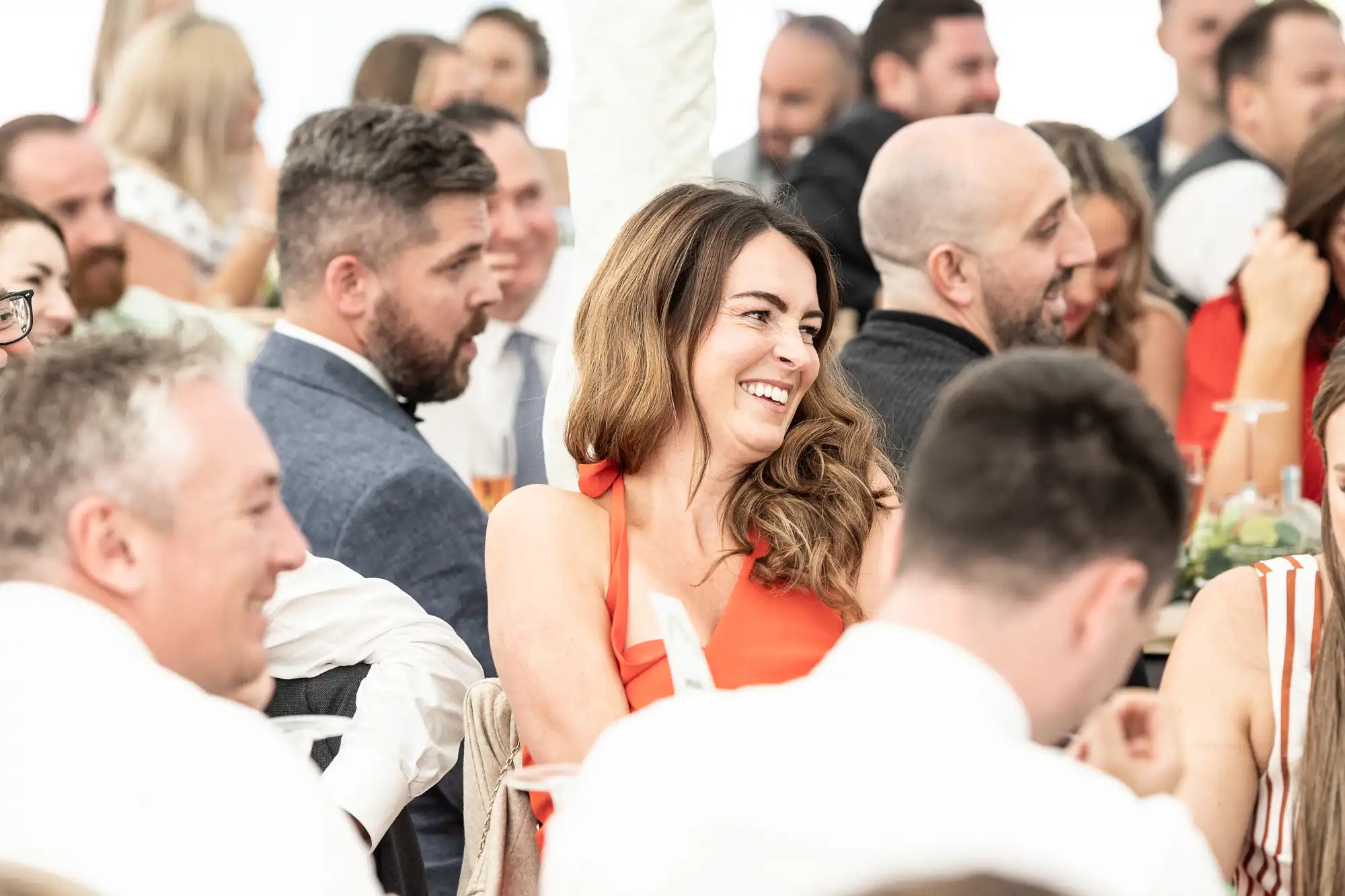 A woman in an orange dress sits smiling at a lively indoor gathering, surrounded by other attendees engaged in conversation.