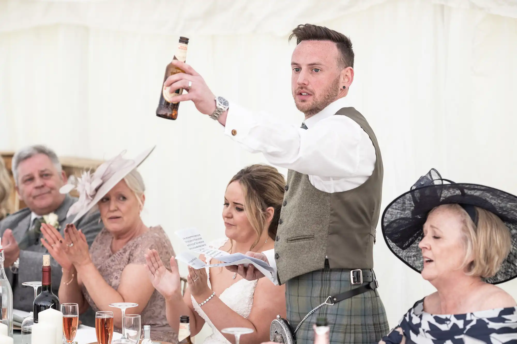 A man in a vest raises a bottle while speaking to a seated audience, including a bride and others dressed formally. Glasses and bottles are on the table.