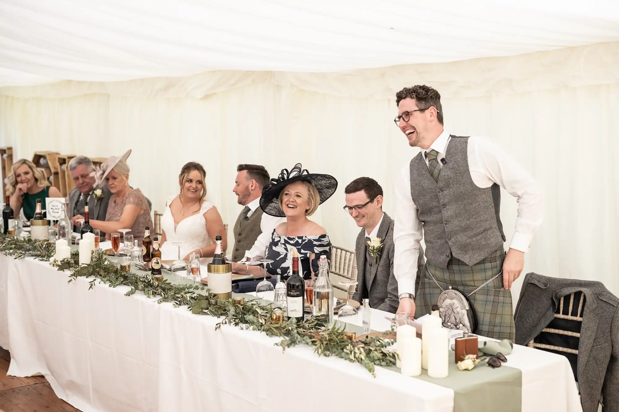 A group of people, dressed in formal attire, are seated at a long table during a wedding reception. One man, standing and smiling, appears to be giving a speech while others listen and smile.