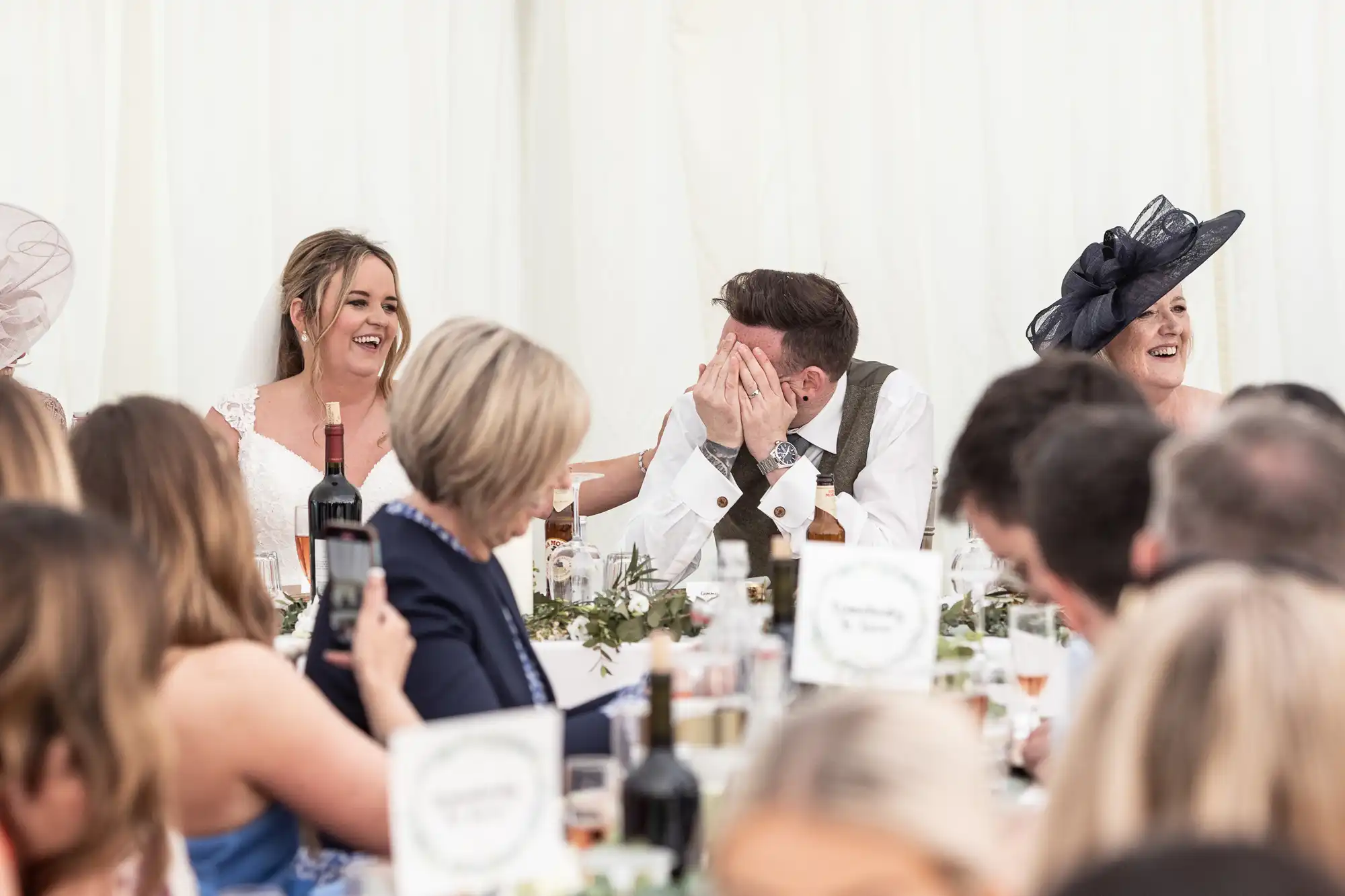 A bride laughs while seated at a table with a groom who covers his face with his hands. Guests around them are engaged and smiling.