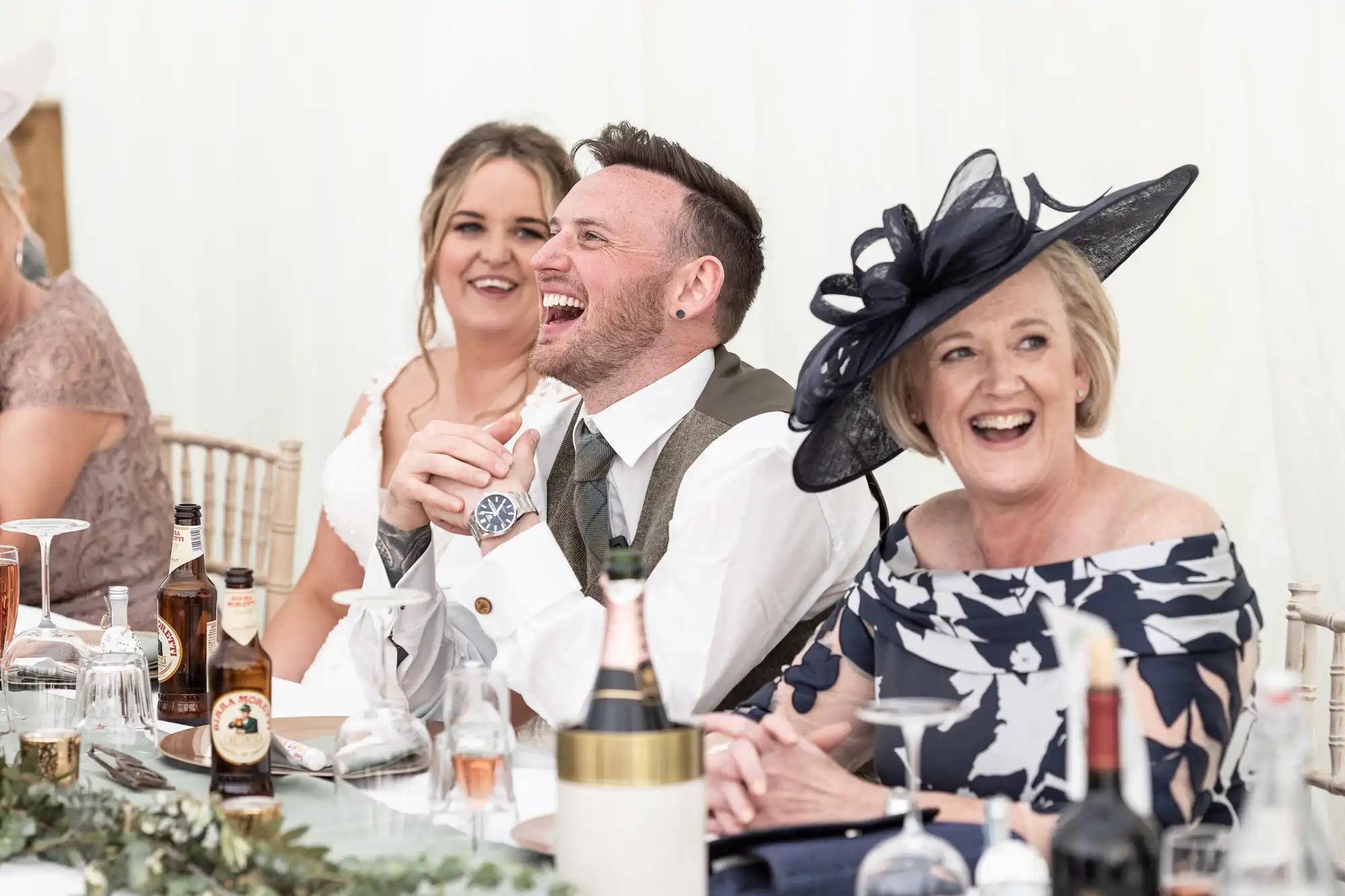 Three people seated at a table, laughing and smiling at an event. Bottles and glasses are on the table, with greenery used as table decoration.