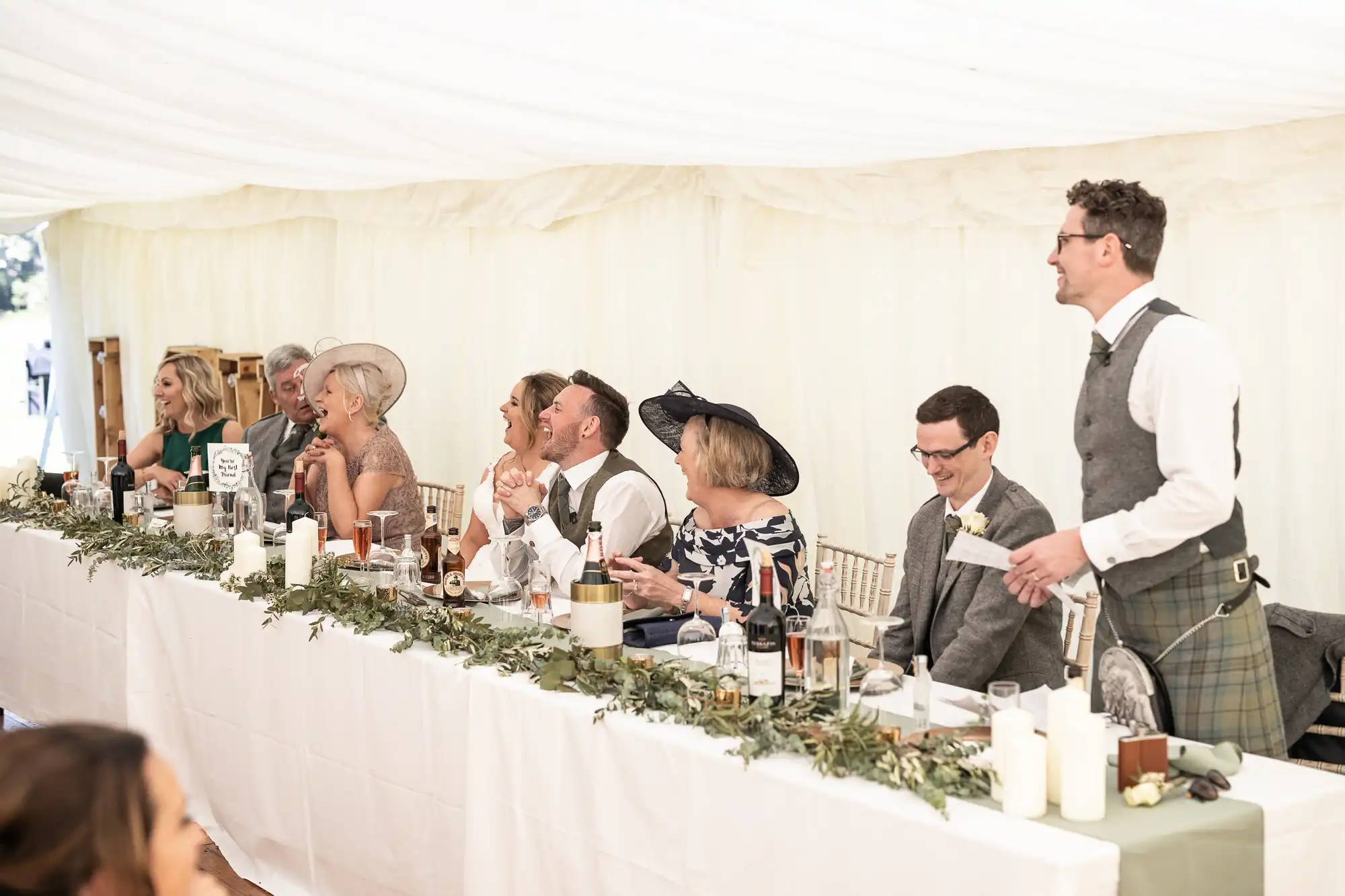 A group of people in formal attire sit at a long, white-clothed table under a tent, while a man in a kilt stands and speaks. The table is adorned with candles and greenery.