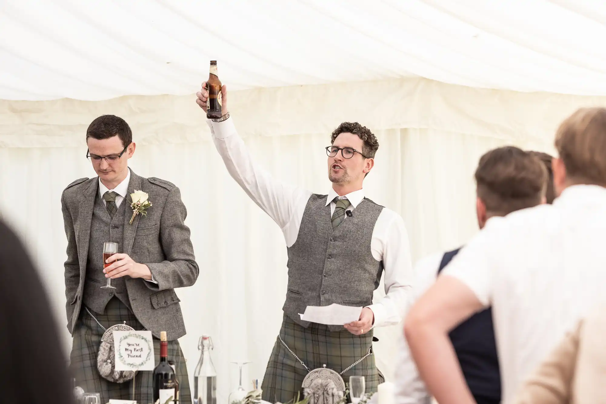 Two men in formal attire give speeches at a wedding reception, one holding up a beer bottle. Guests in the foreground listen attentively.