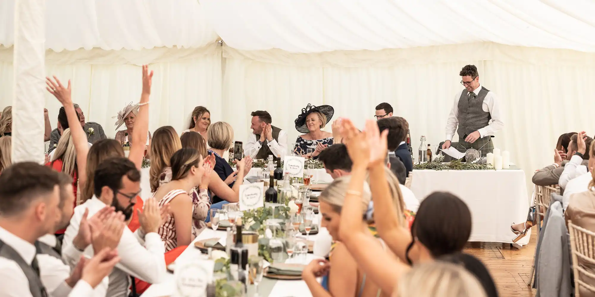 A man stands to give a speech in a decorated tent filled with seated guests applauding and clapping. The tables are set with dinnerware and centerpieces.