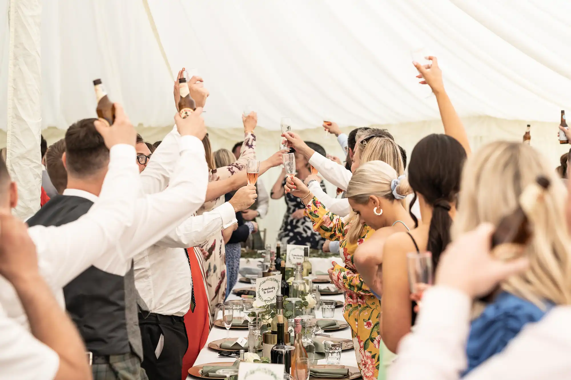 A group of people inside a tent raising glasses and bottles in a toast, seated around a long, decorated table.