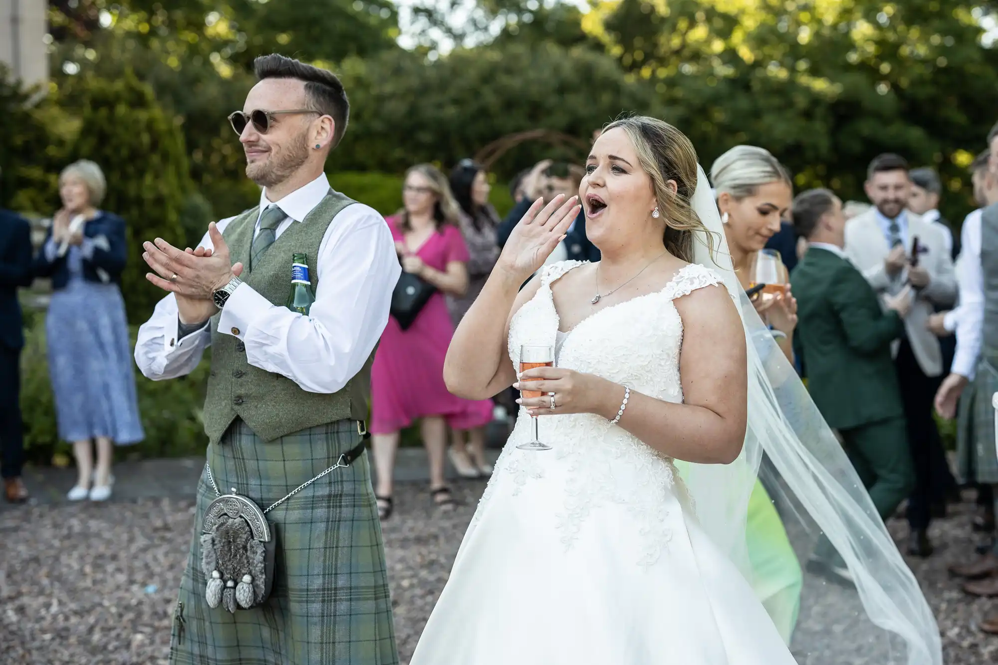 A bride and groom stand together in a garden. The groom, wearing a kilt, claps while the bride, in a white gown, holds a drink and appears to be yawning or surprised. Guests are in the background.