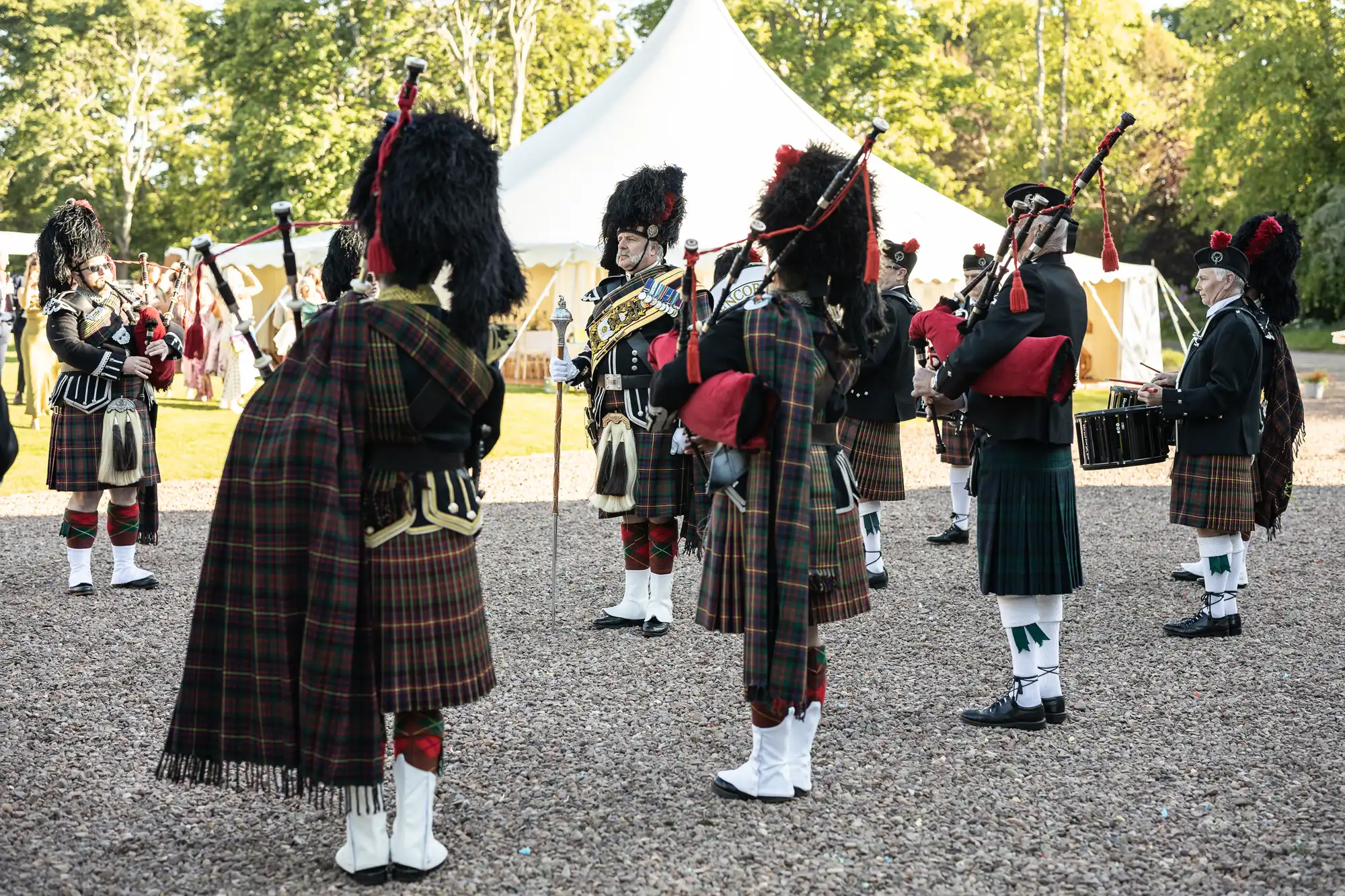 A group of people in traditional Scottish attire, including kilts and feathered caps, play bagpipes and drums in an outdoor setting with a white tent in the background.