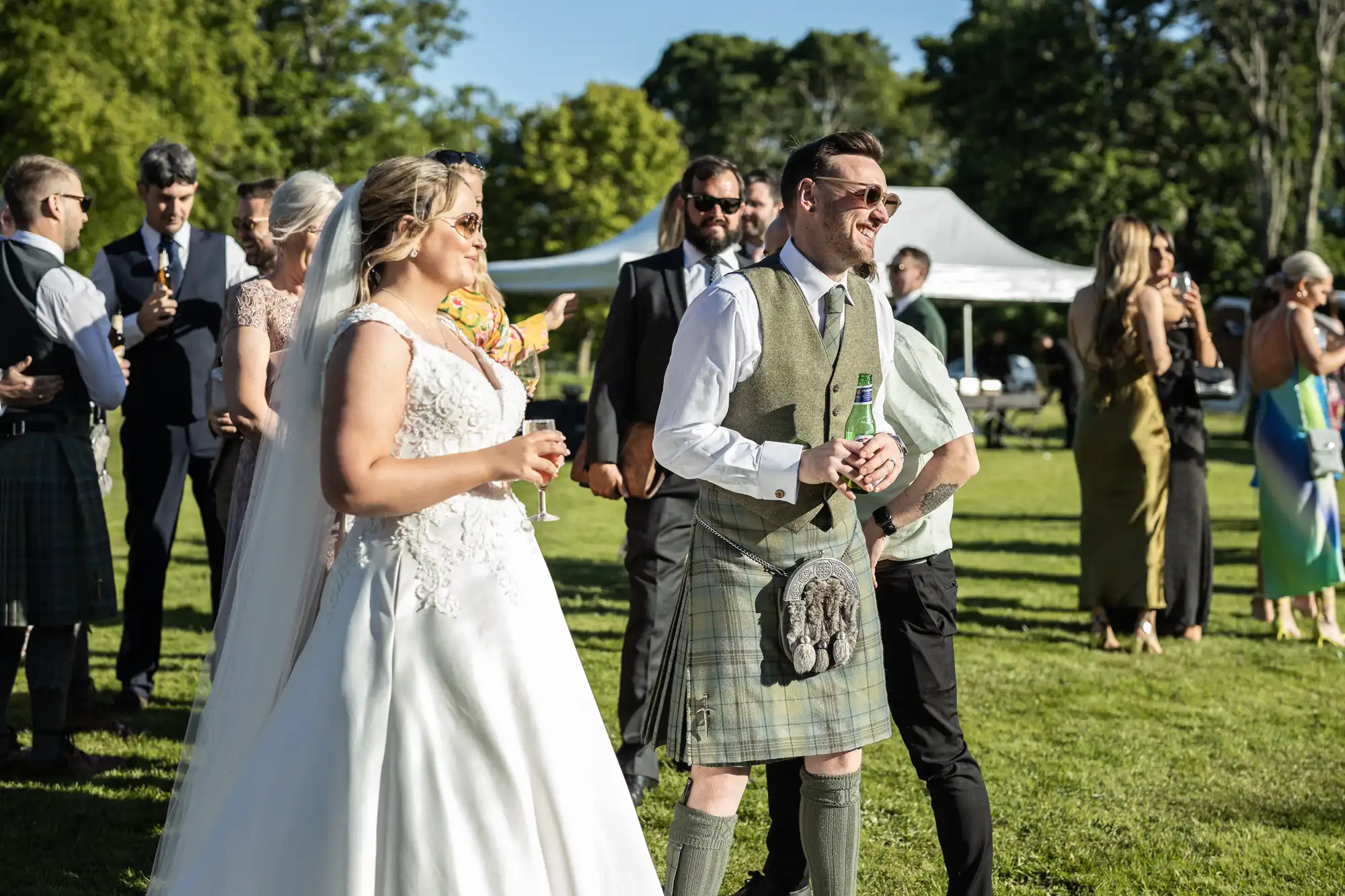 A bride in a white gown and a groom in traditional Scottish attire stand outdoors among guests at a sunny wedding reception.