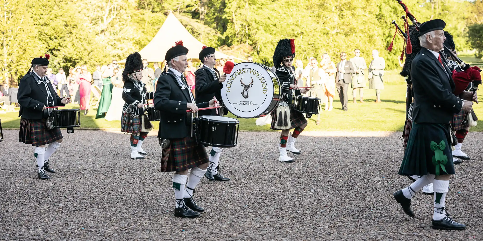 A marching band in traditional Scottish attire performs outdoors, with drummers and bagpipers playing. A drum with "The Stonehouse Pipe Band" logo is visible. Spectators watch in the background.