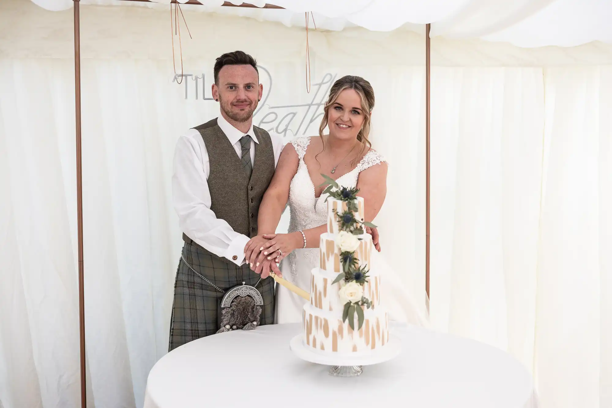 A couple stands together, cutting a tiered wedding cake adorned with flowers. The groom wears a vest and a kilt, while the bride is in a white dress with lace details.