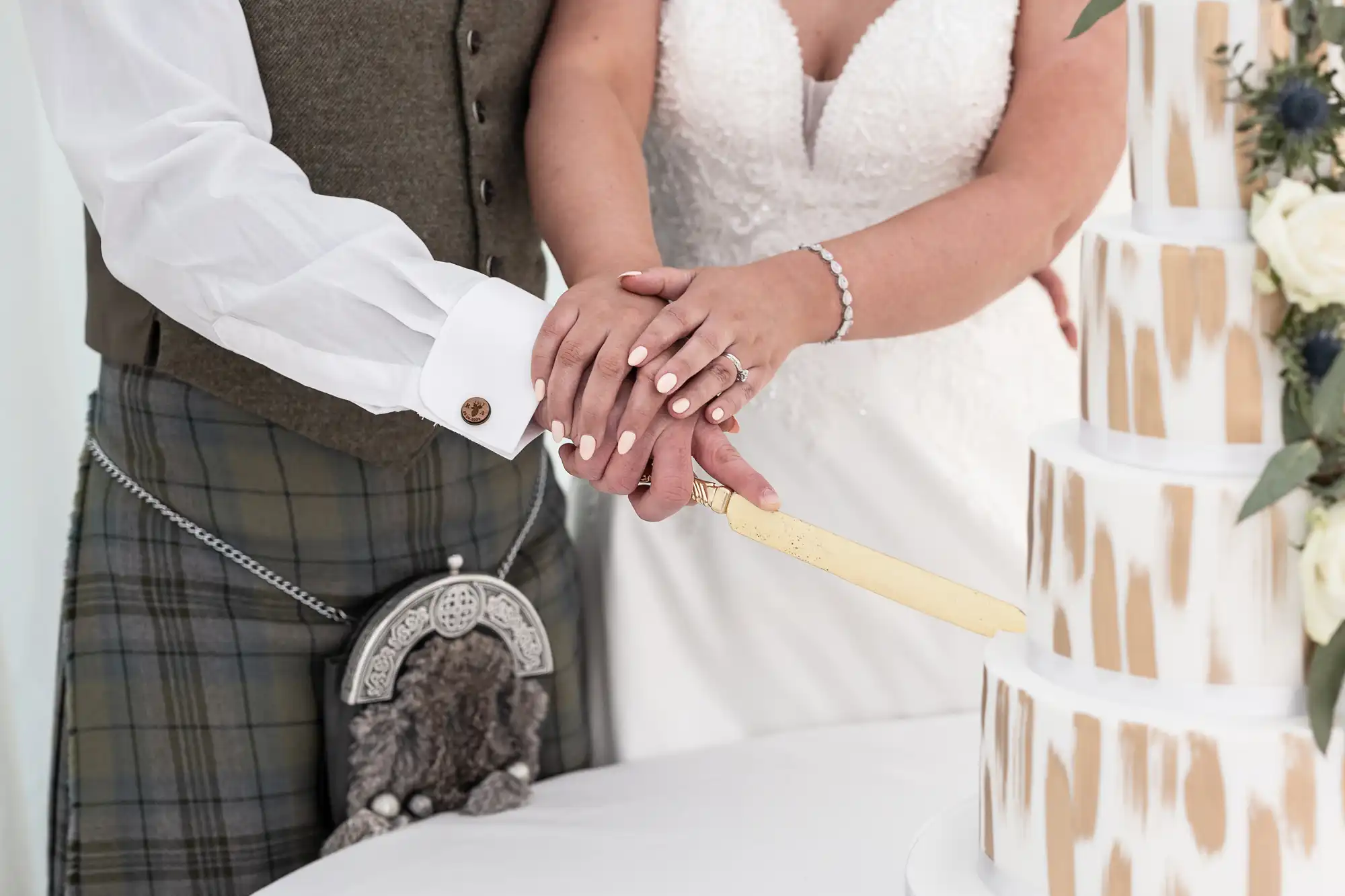 A couple cuts a tiered wedding cake together, with their hands overlapped on the knife. The groom wears a kilt and the bride is in a white dress.