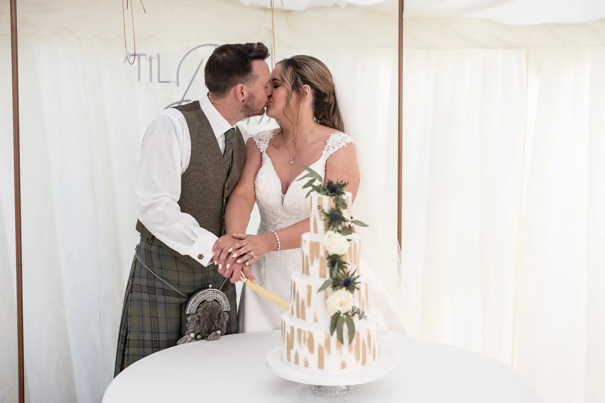 A bride and groom kiss while cutting a three-tiered wedding cake adorned with white roses and greenery. The groom wears a tartan kilt and vest, and the bride wears a white lace wedding dress.