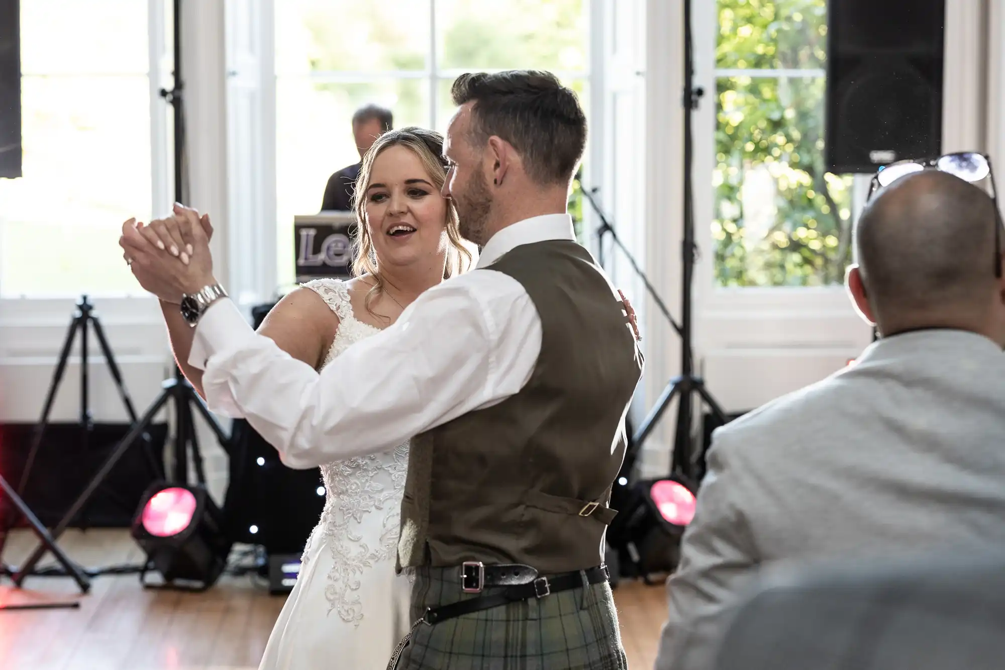 A bride and groom dance together in a brightly lit room with a band setup in the background. The groom wears a plaid vest and kilt, and the bride is in a white wedding dress.